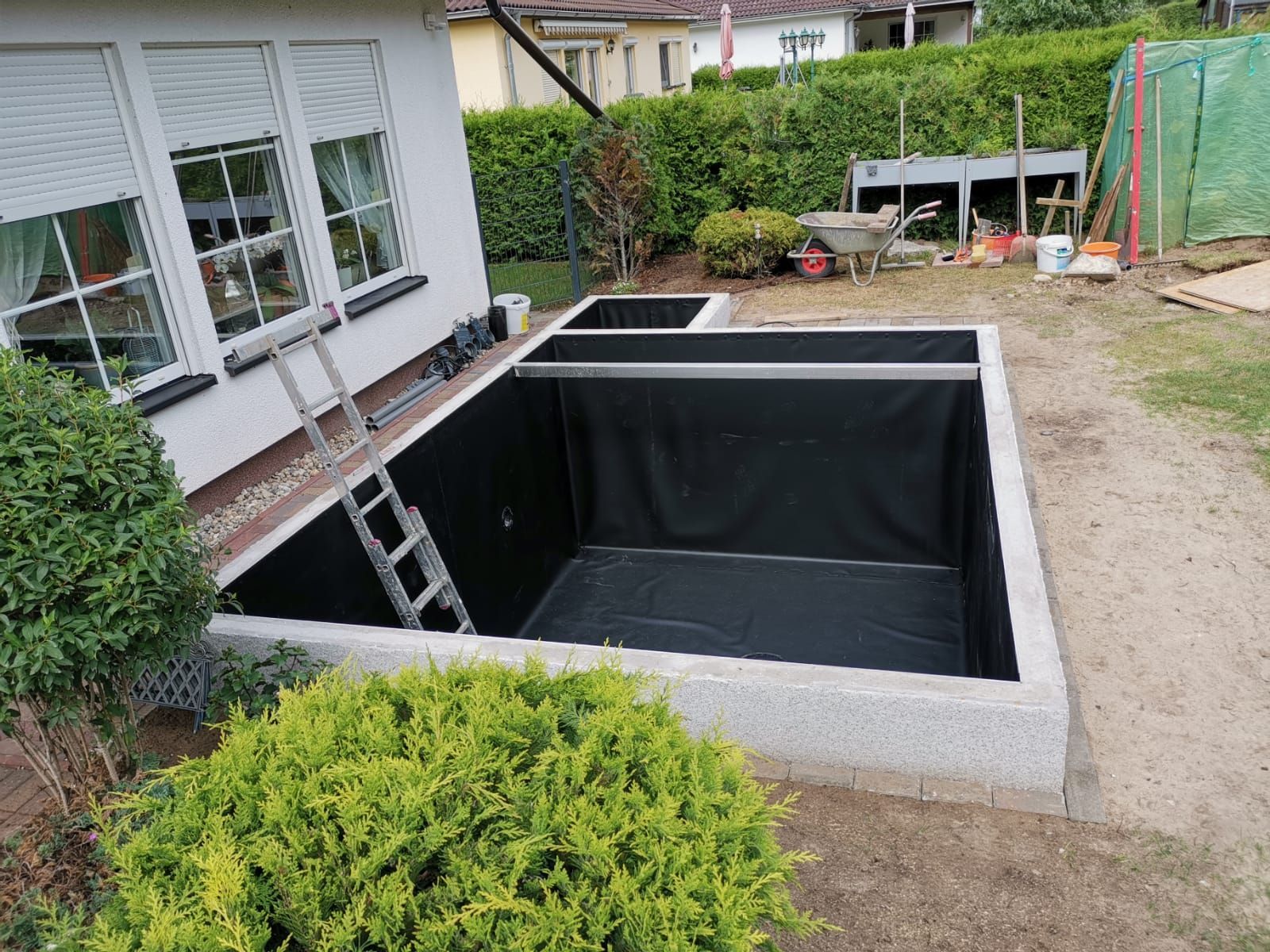 Mit Betonwänden und schwarzem Material ausgekleideter Außenteich in der Nähe eines Hauses mit Fenstern;