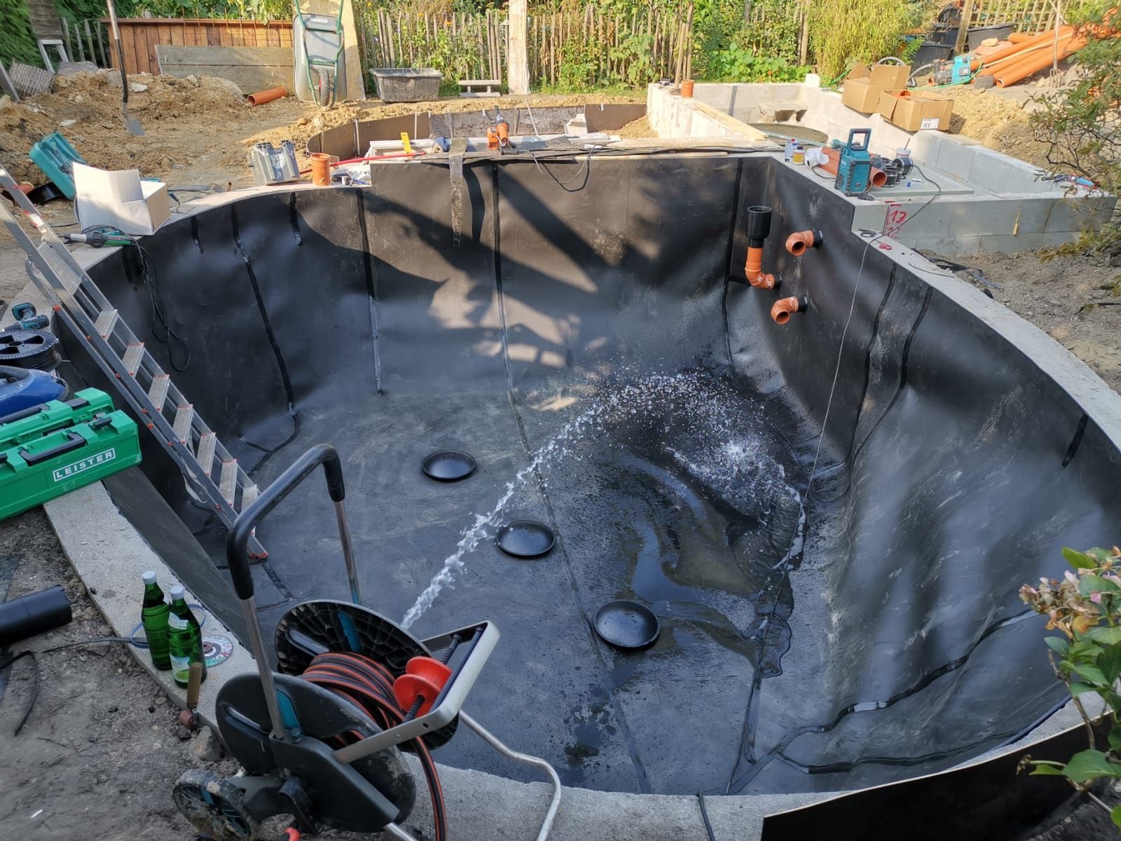 Bau eines kleinen Pools mit schwarzer Auskleidung, umgeben von Beton, im Außenbereich. Wasser spritzt aus einem Schlauch.