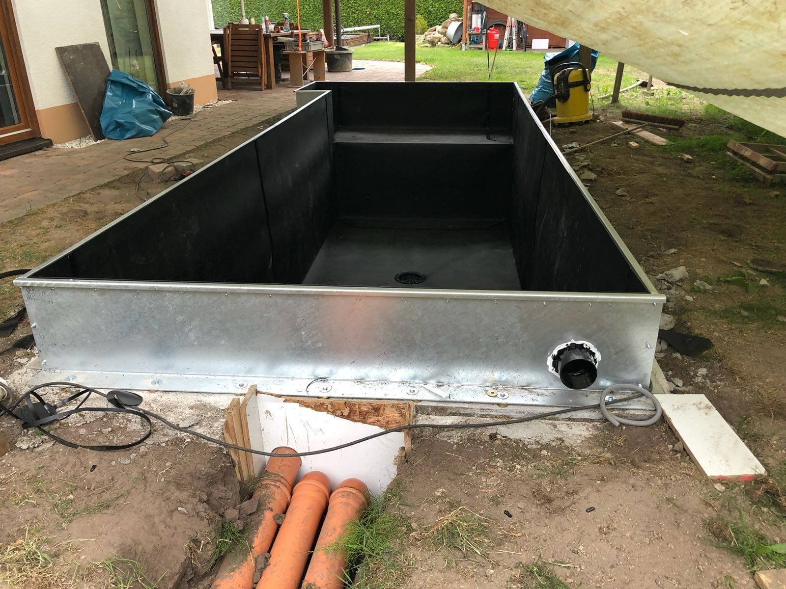 Im Außenbereich entsteht ein rechteckiger Pool mit schwarzer Auskleidung. Darunter sind orangefarbene Abflussrohre zu sehen.