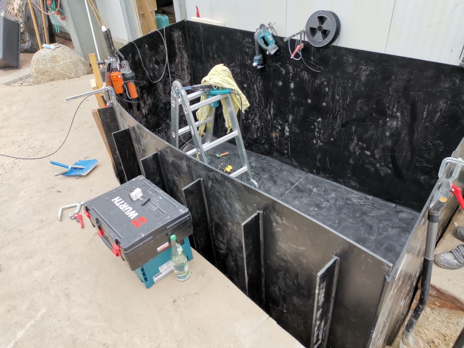 Schwarze rechteckige Struktur mit Leiter im Inneren. Werkzeuge und Geräte umgeben sie.