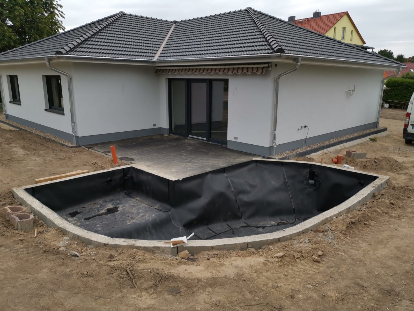 Ein teilweise angelegter Gartenteich vor einem weißen Haus mit dunklem Dach und Glasschiebetüren. Die Teichfolie ist schwarz.