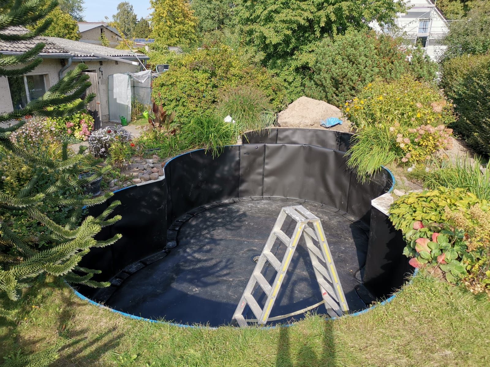 Ein leerer, schwarz umrandeter Teich im Bau, mit einer Leiter darin. Umgeben von grünem Gras, Bäumen und einem Garten.