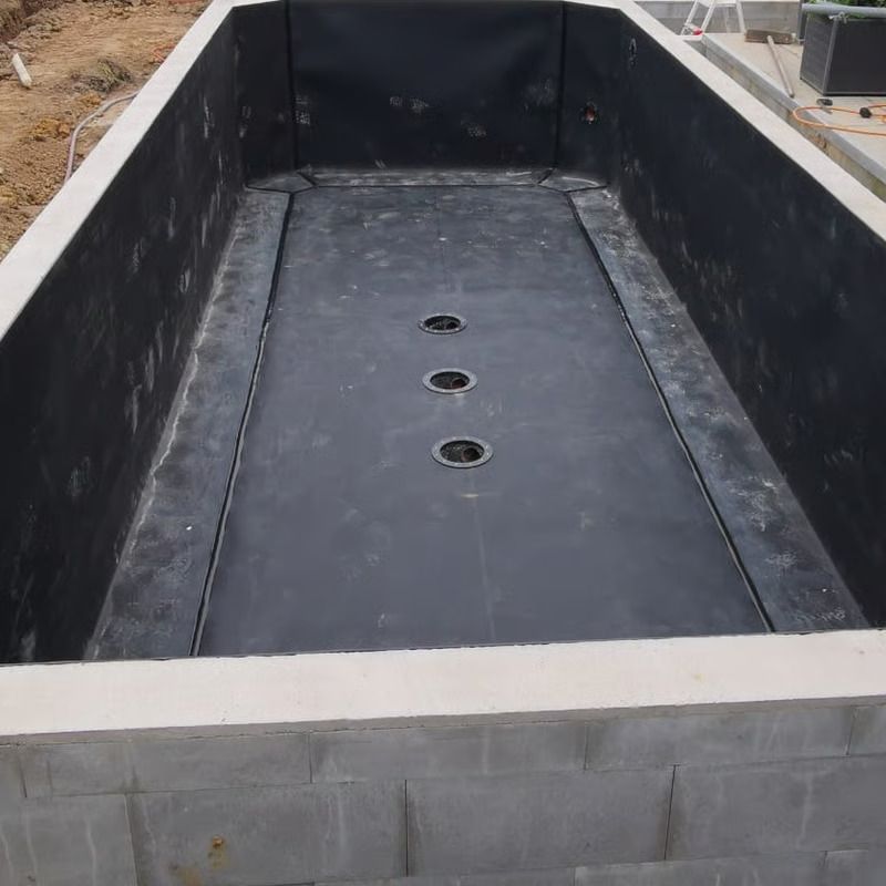 Rechteckiger Betonpool im Bau, mit schwarzem Material ausgekleidet. Am Boden sind drei Abflüsse sichtbar.