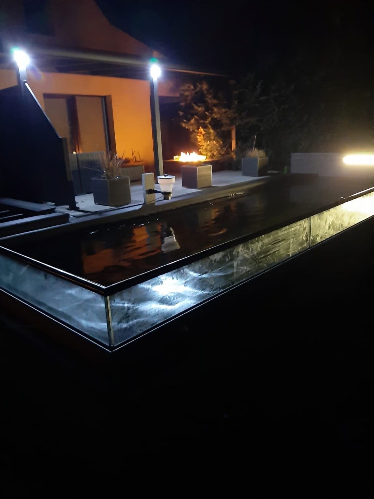 Nachtansicht eines rechteckigen Pools mit Beleuchtung, einer Terrasse mit Sitzgelegenheiten und einer Feuerstelle.