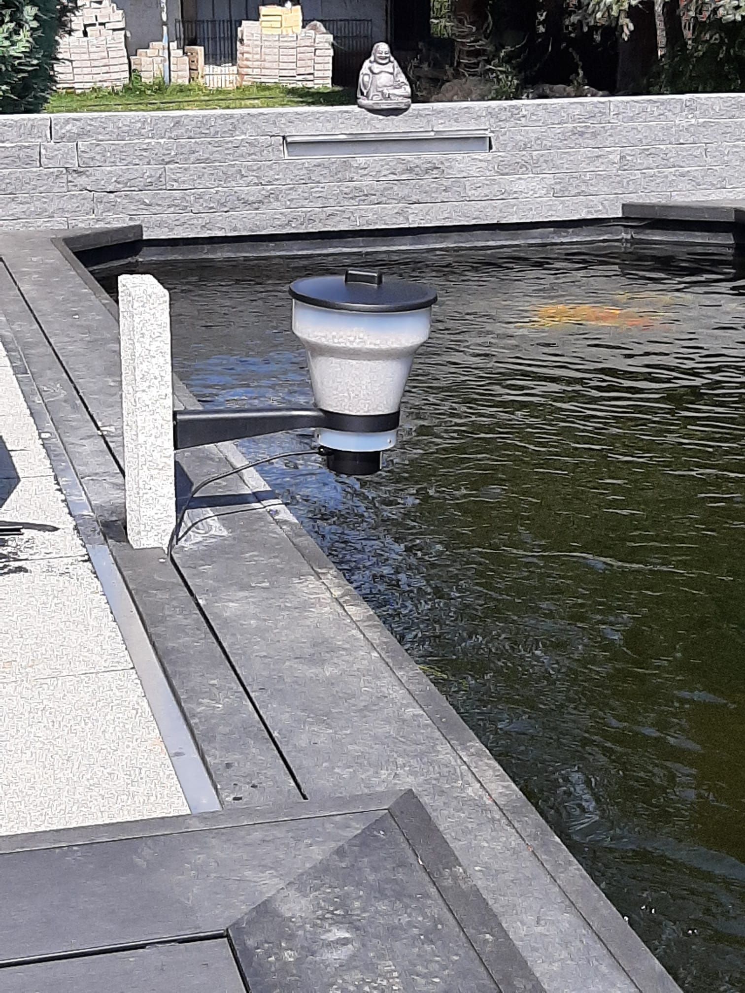 Brunnen in einem Teich mit grauen Steinstufen und einer dekorativen Skulptur im Hintergrund. Wasser fließt aus dem Brunnen.