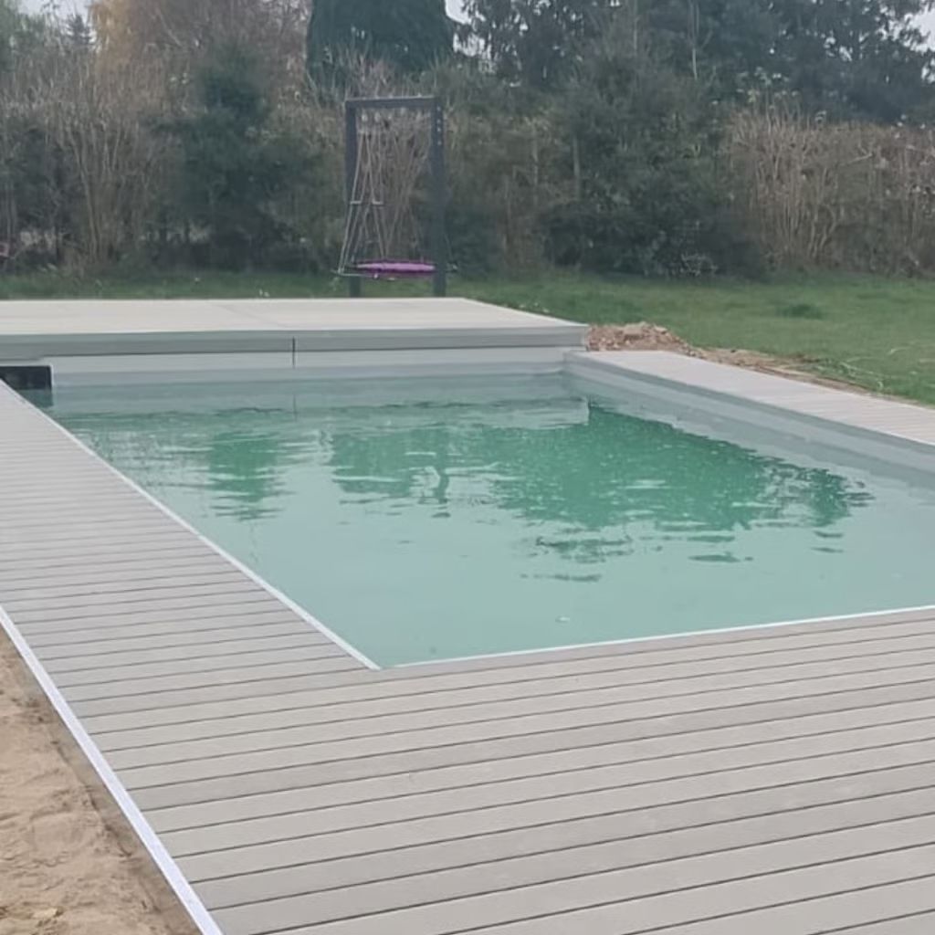 Pool mit hellgrauer Terrasse, umgeben von Gras und Bäumen, mit einer Schaukel im Hintergrund.