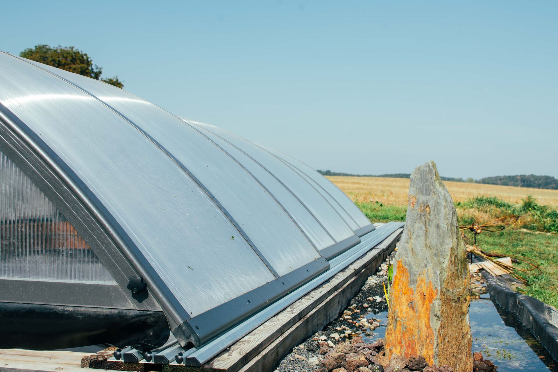 Ein teilweise offenes Gewächshaus mit gebogenen, durchscheinenden Dachpaneelen, in der Nähe eines Feldes unter blauem Himmel.