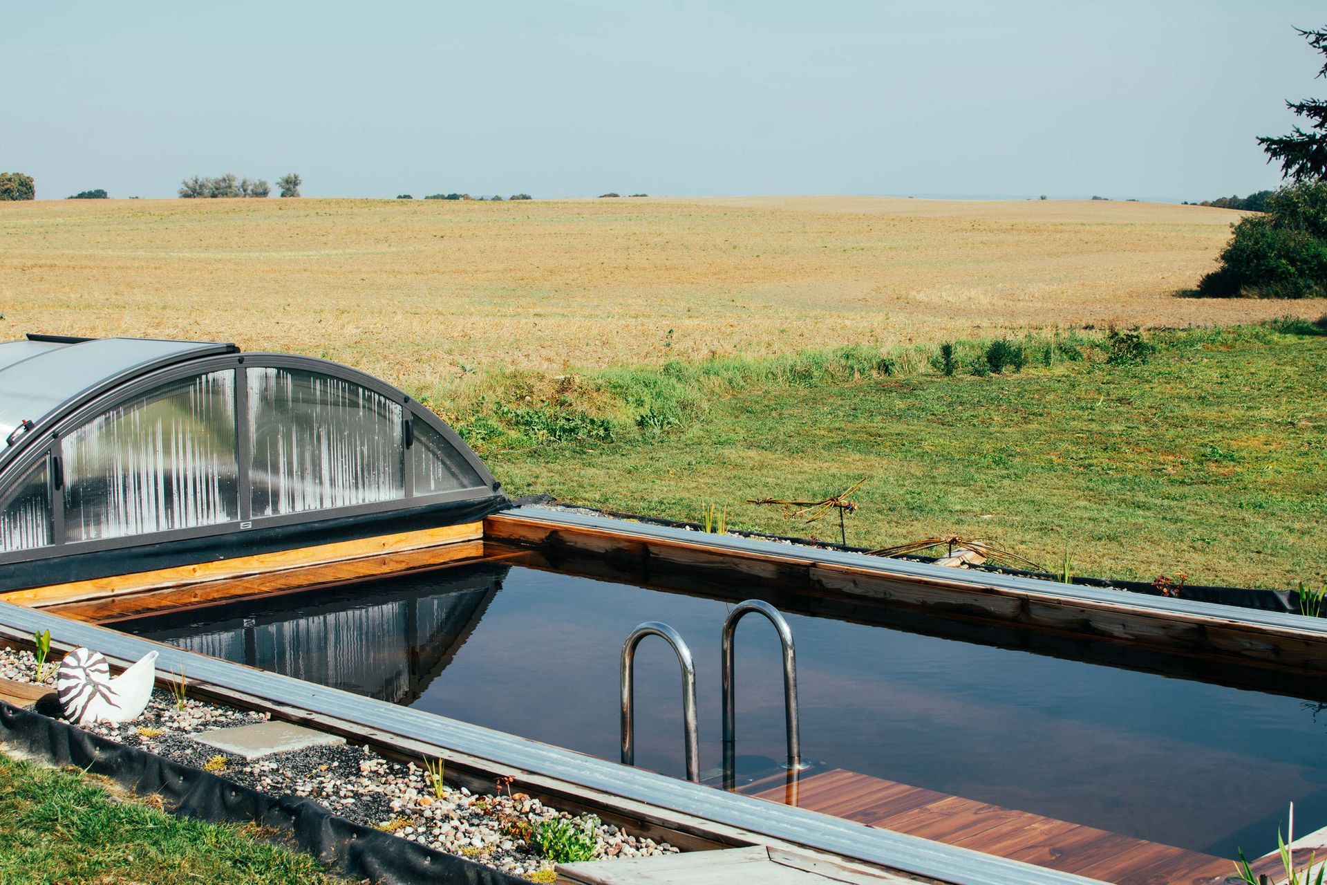 Swimmingpool mit Abdeckung, Leiter und Holzdeck, eingebettet in eine grüne Wiese mit einem Feld im Hintergrund.