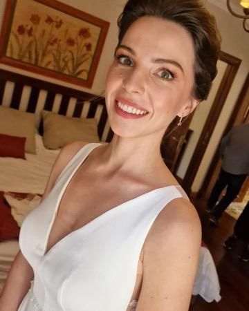 Mujer con vestido blanco sonríe, en el interior. Dormitorio con cabecero de madera, cuadro y persona al fondo.