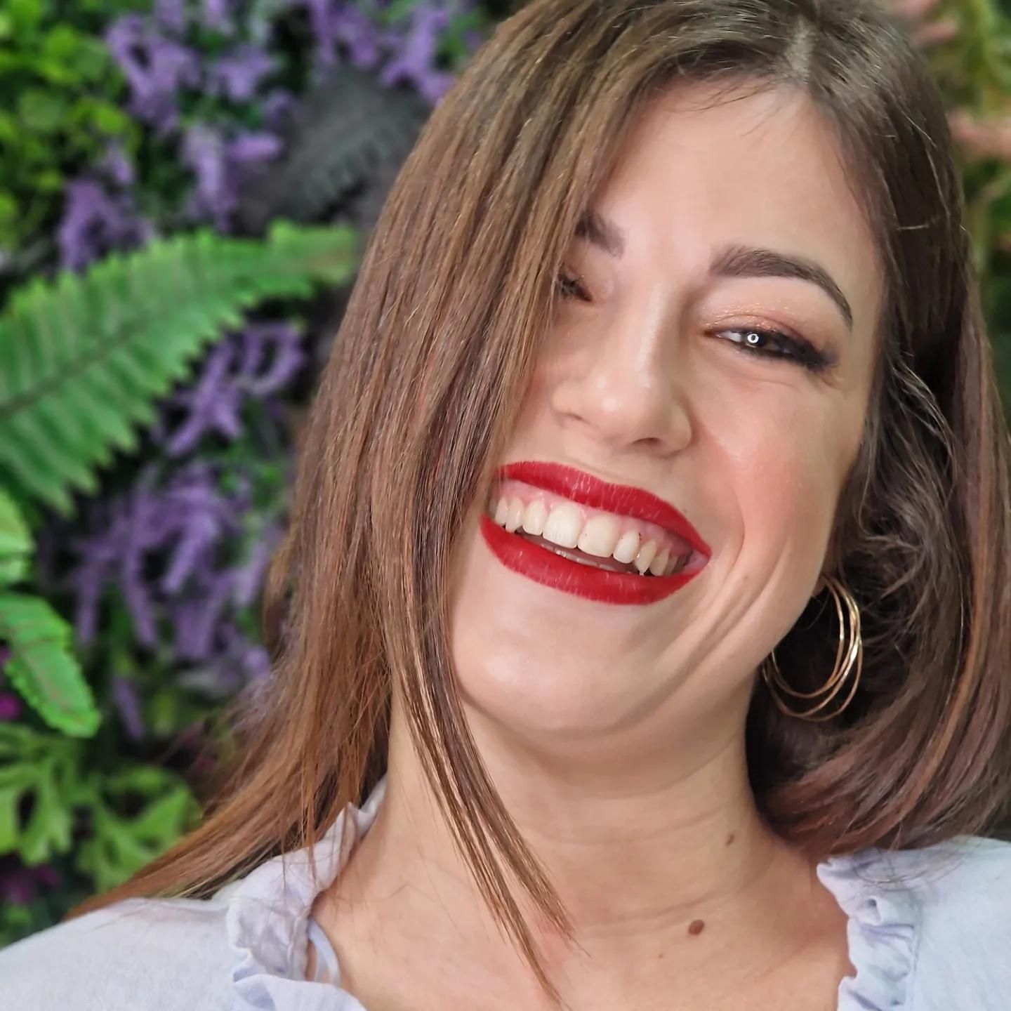Mujer de cabello castaño sonriendo, con lápiz labial rojo y aretes de aro dorados, frente a vegetación.