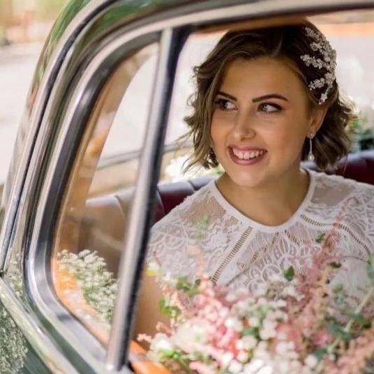 Novia en un coche clásico, sonriente, con un vestido blanco de encaje y un adorno floral en el pelo, sosteniendo flores.