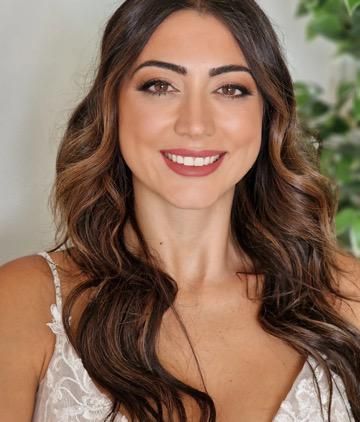 Mujer de cabello castaño largo y ondulado, vestida con un vestido blanco, sonriendo con maquillaje de ojos oscuro