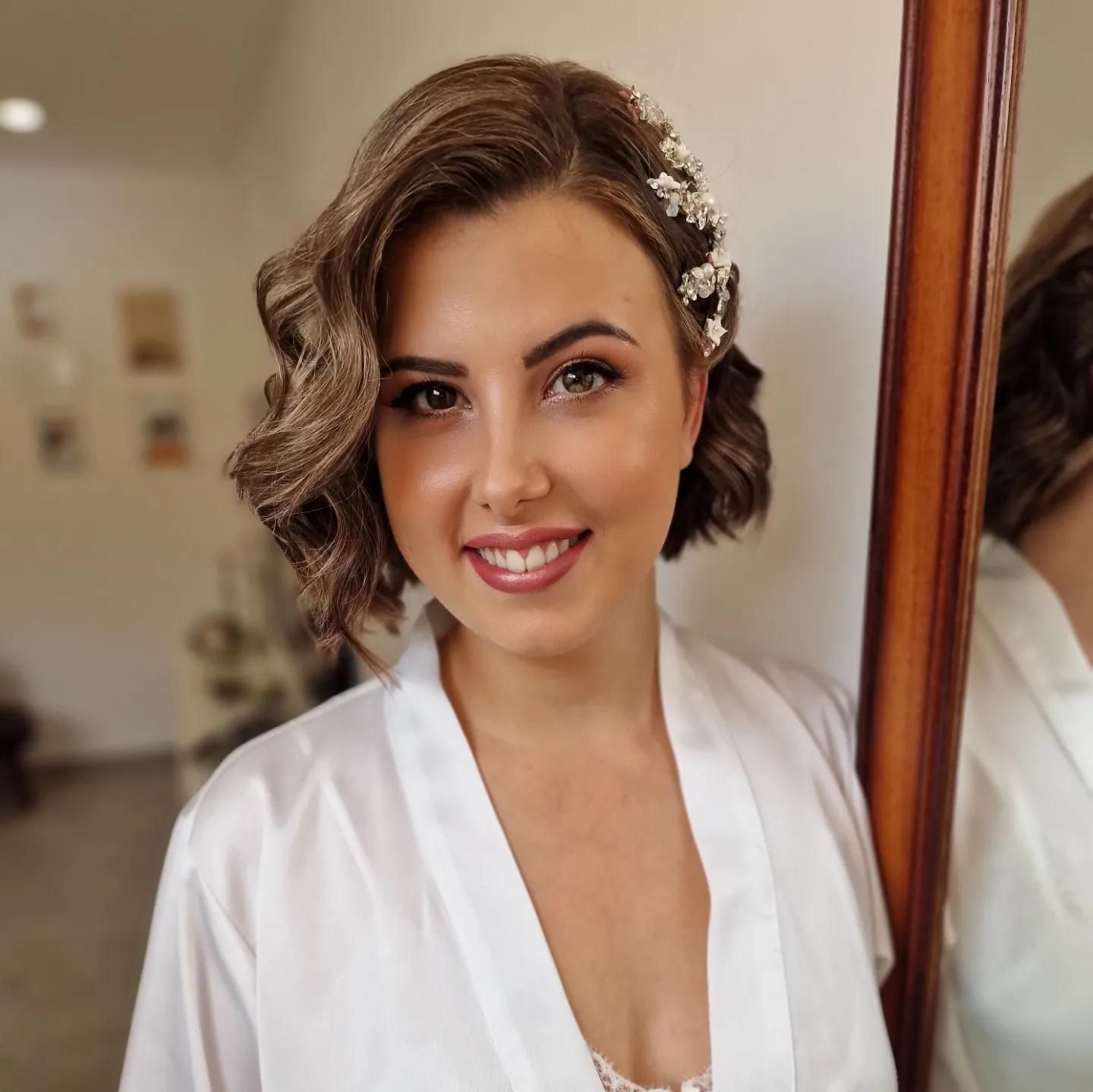 Mujer con el pelo corto y ondulado y maquillaje de novia, vestida con una bata blanca, sonriendo.