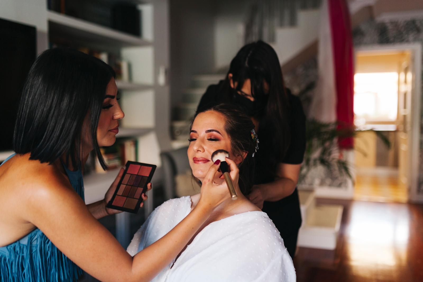 Una maquilladora aplica colorete a una mujer con bata blanca. Otra maquilladora le peina el cabello en el interior.