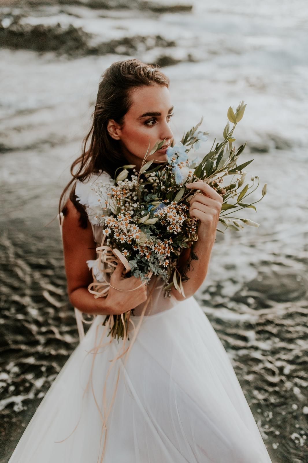 Mujer con vestido blanco oliendo un ramo de flores en una playa con olas del océano.