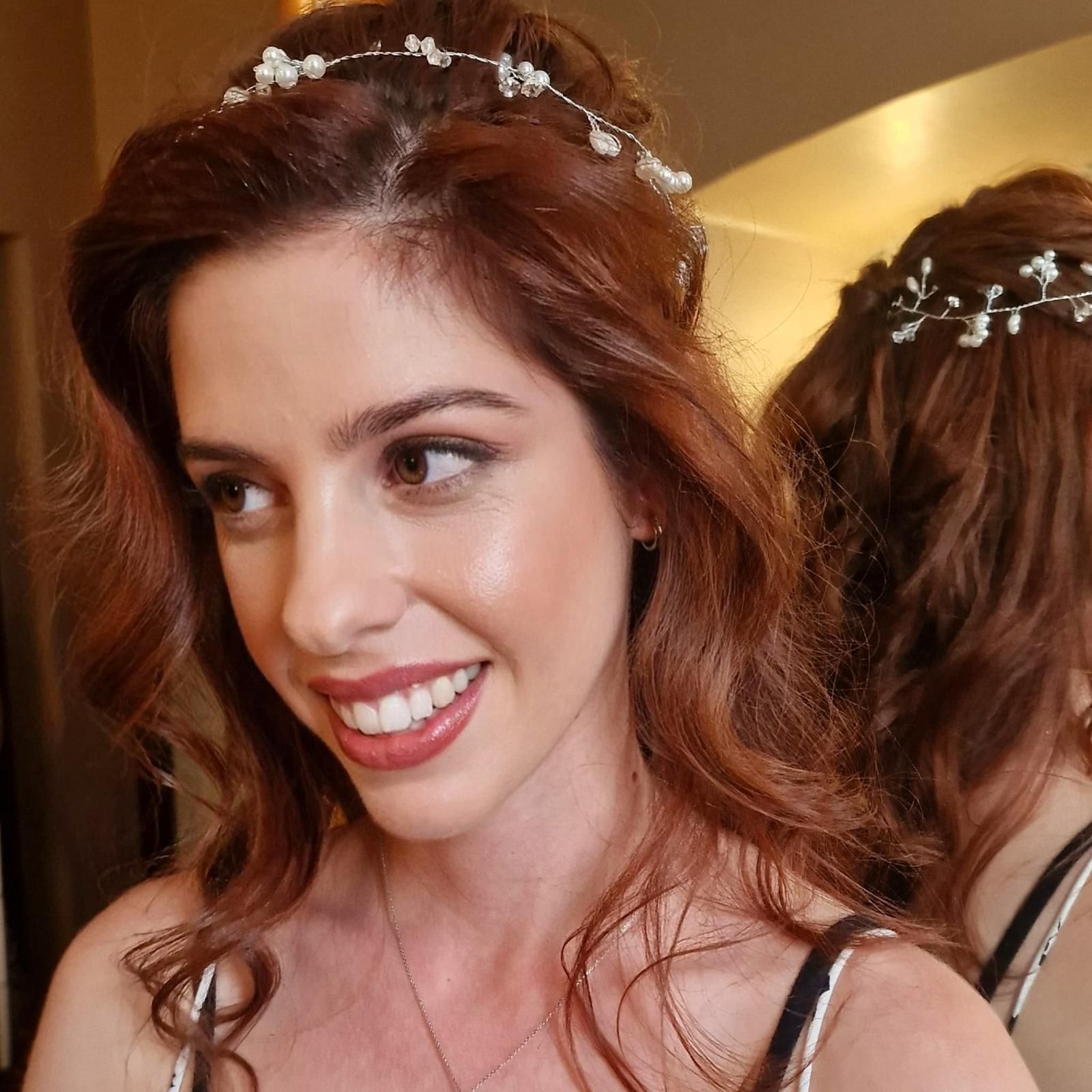 Una mujer de cabello castaño rojizo sonríe, luciendo un adorno de perlas en el cabello. Un espejo refleja su espalda.