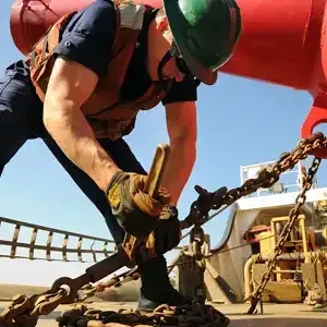 Ein Mann mit Schutzhelm und Handschuhen arbeitet an einer Kette auf einem Boot.