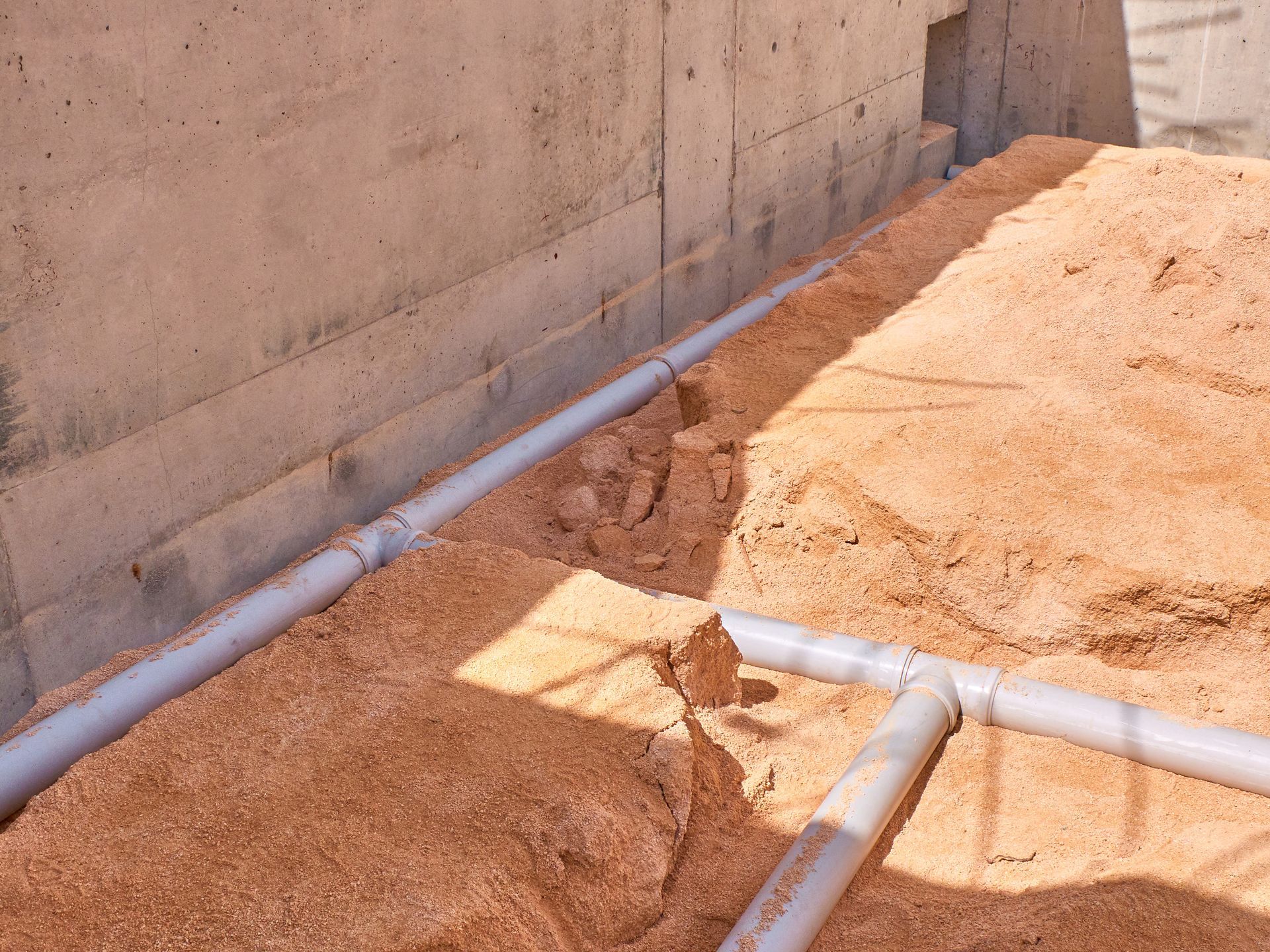 Des tuyaux en PVC gris posés dans du sable brun rougeâtre, le long d'un mur de fondations en béton.