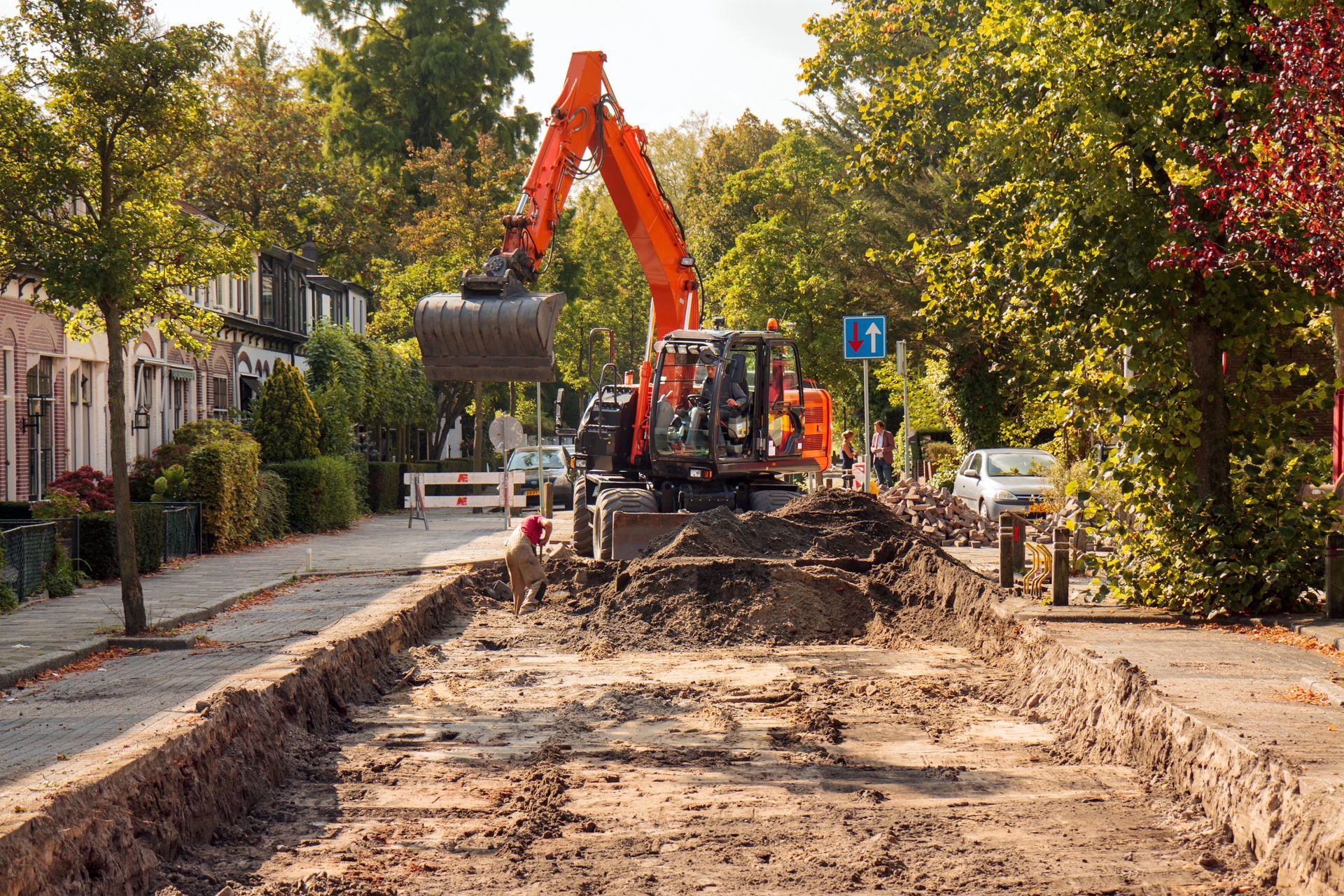 Une excavatrice creuse une tranchée dans une rue résidentielle ; machine orange, terre, arbres, soleil.