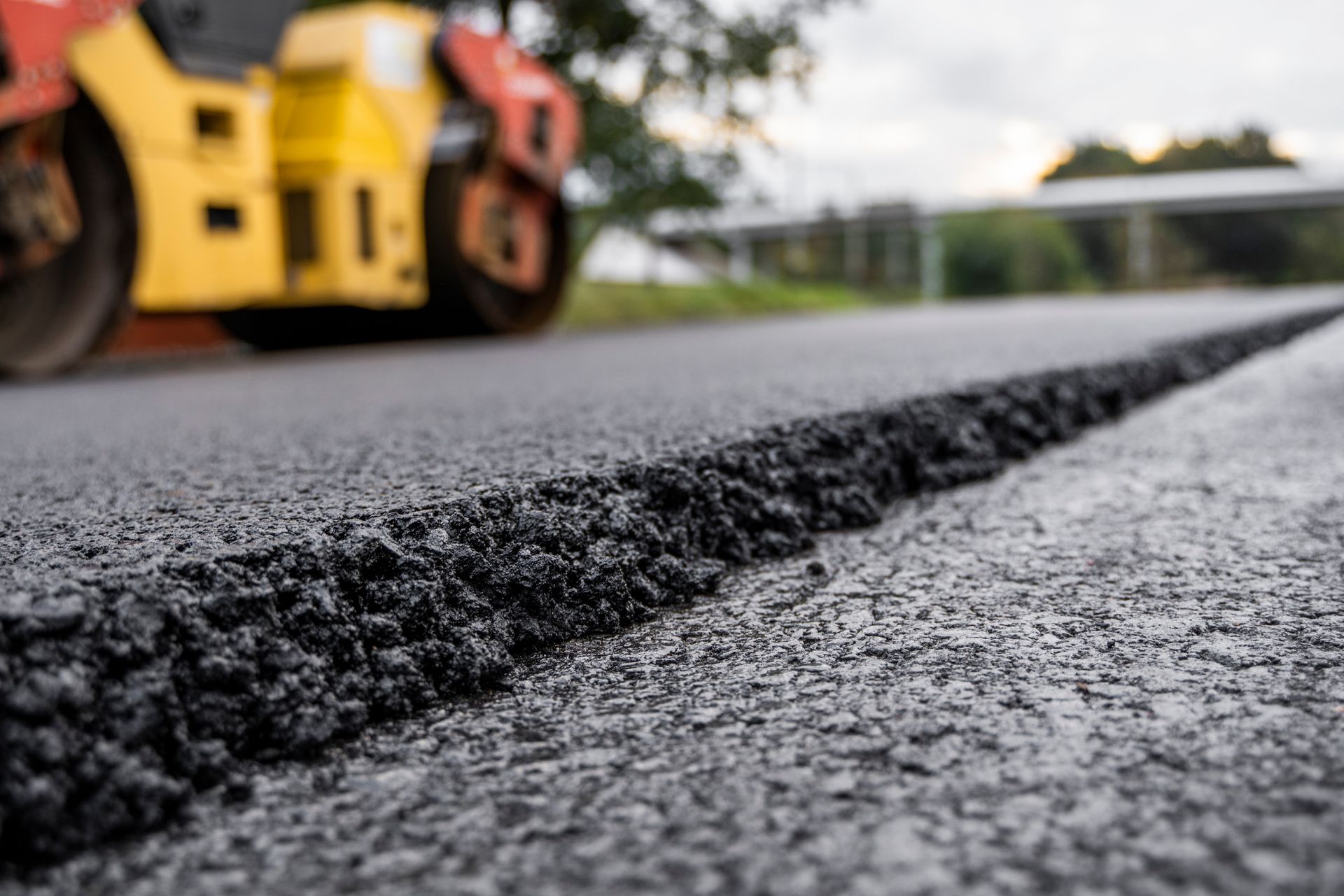 Route asphaltée fraîchement pavée avec un rouleau compresseur jaune et rouge en arrière-plan flou.