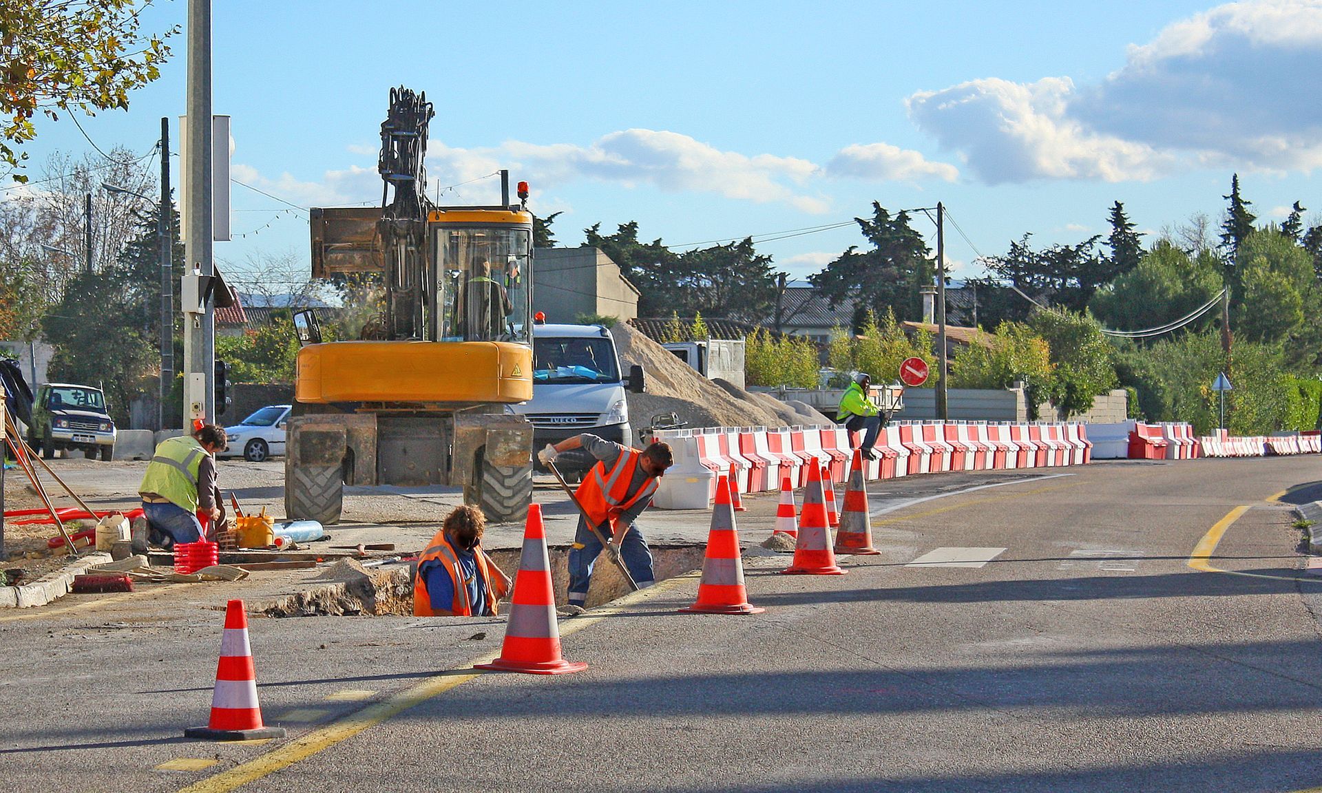 Chantier routier : des ouvriers en gilets orange travaillent près d'une excavatrice, de cônes orange et de barrières sur une route asphaltée sous un ciel bleu.