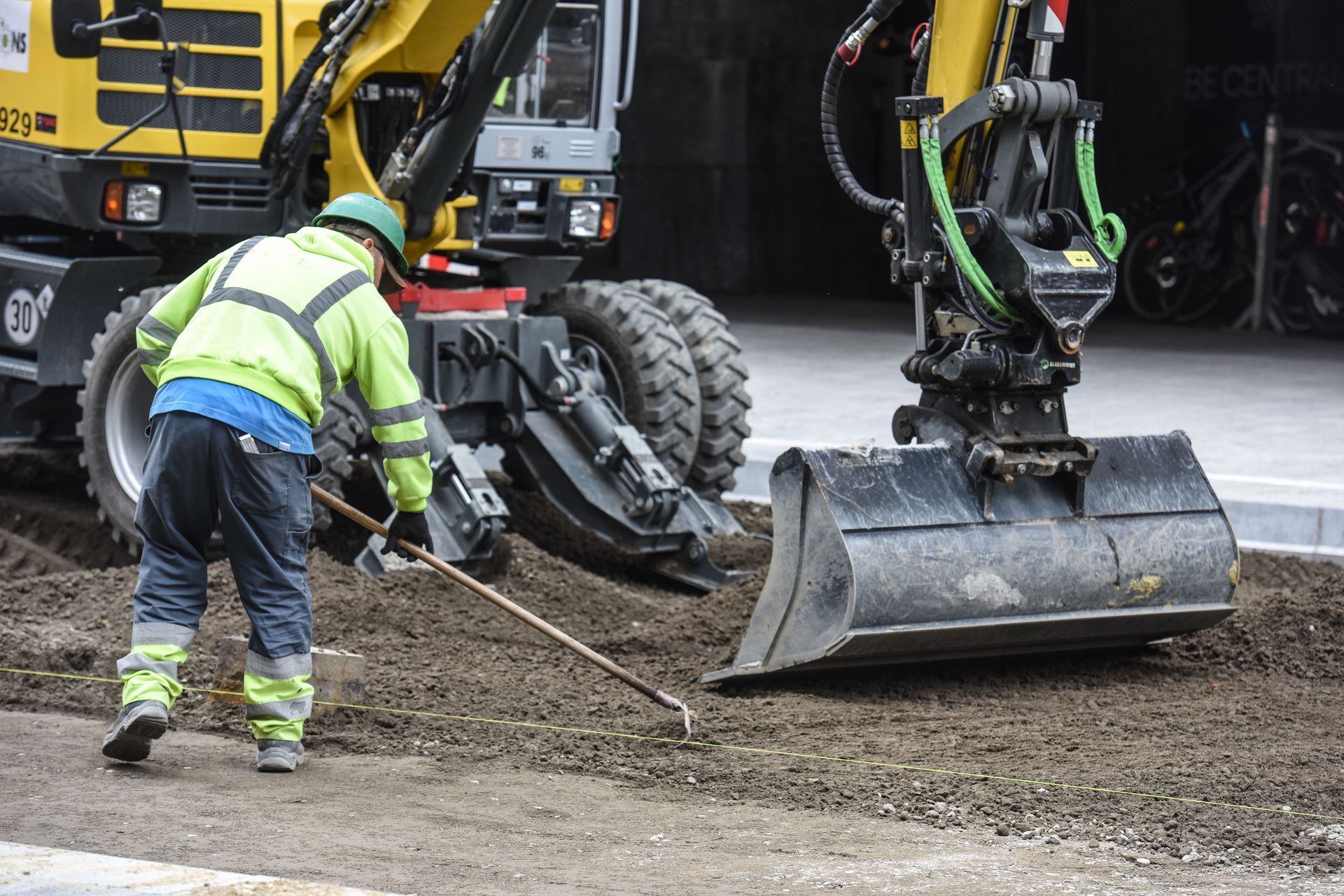 Un ouvrier du bâtiment ratisse du gravier à côté d'une excavatrice. Il porte un gilet de sécurité.