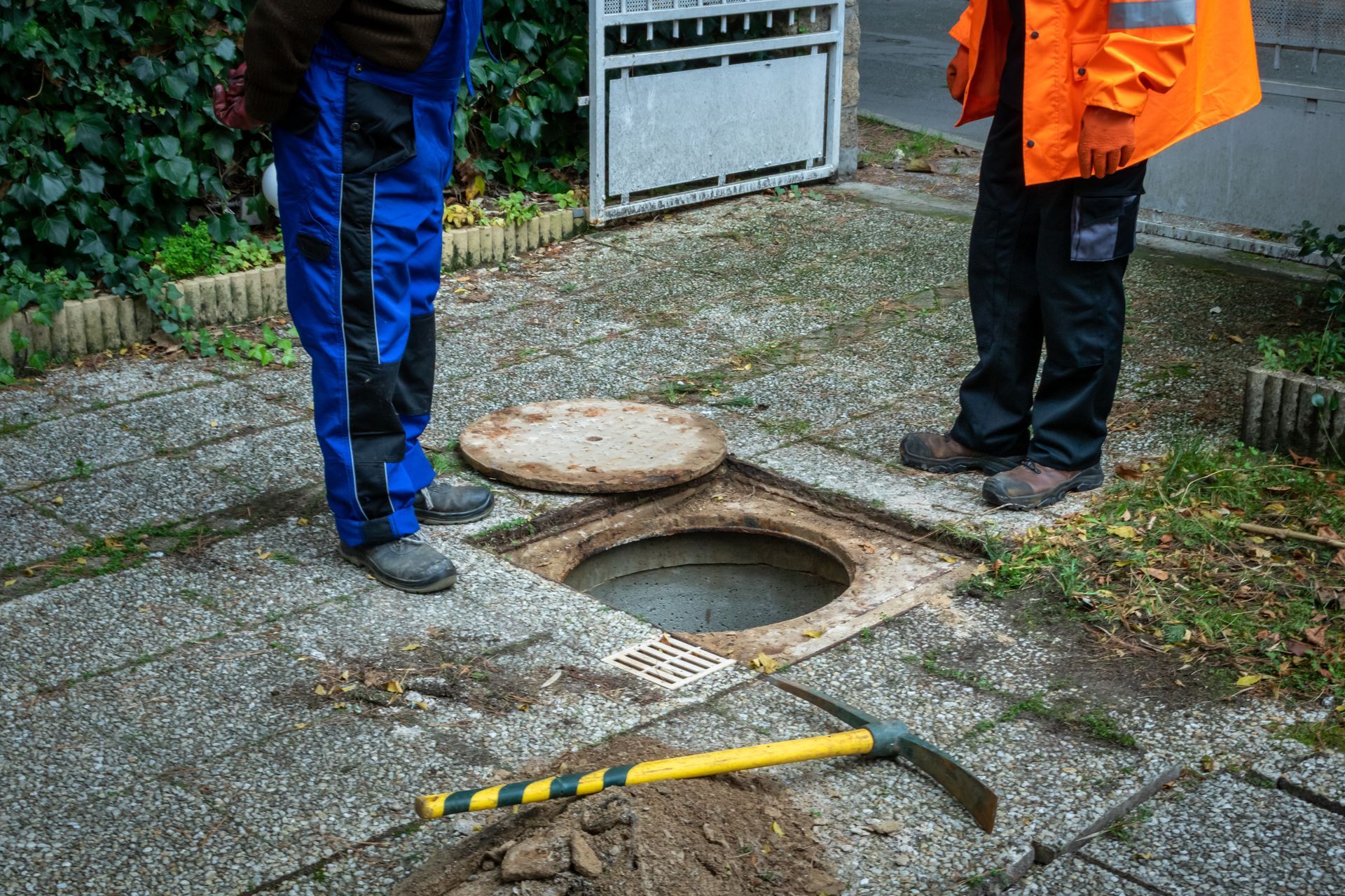 Deux ouvriers se trouvent près d'une bouche d'égout ouverte sur une surface pavée. L'un porte une salopette bleue, l'autre un gilet orange. Une pioche gît à proximité.