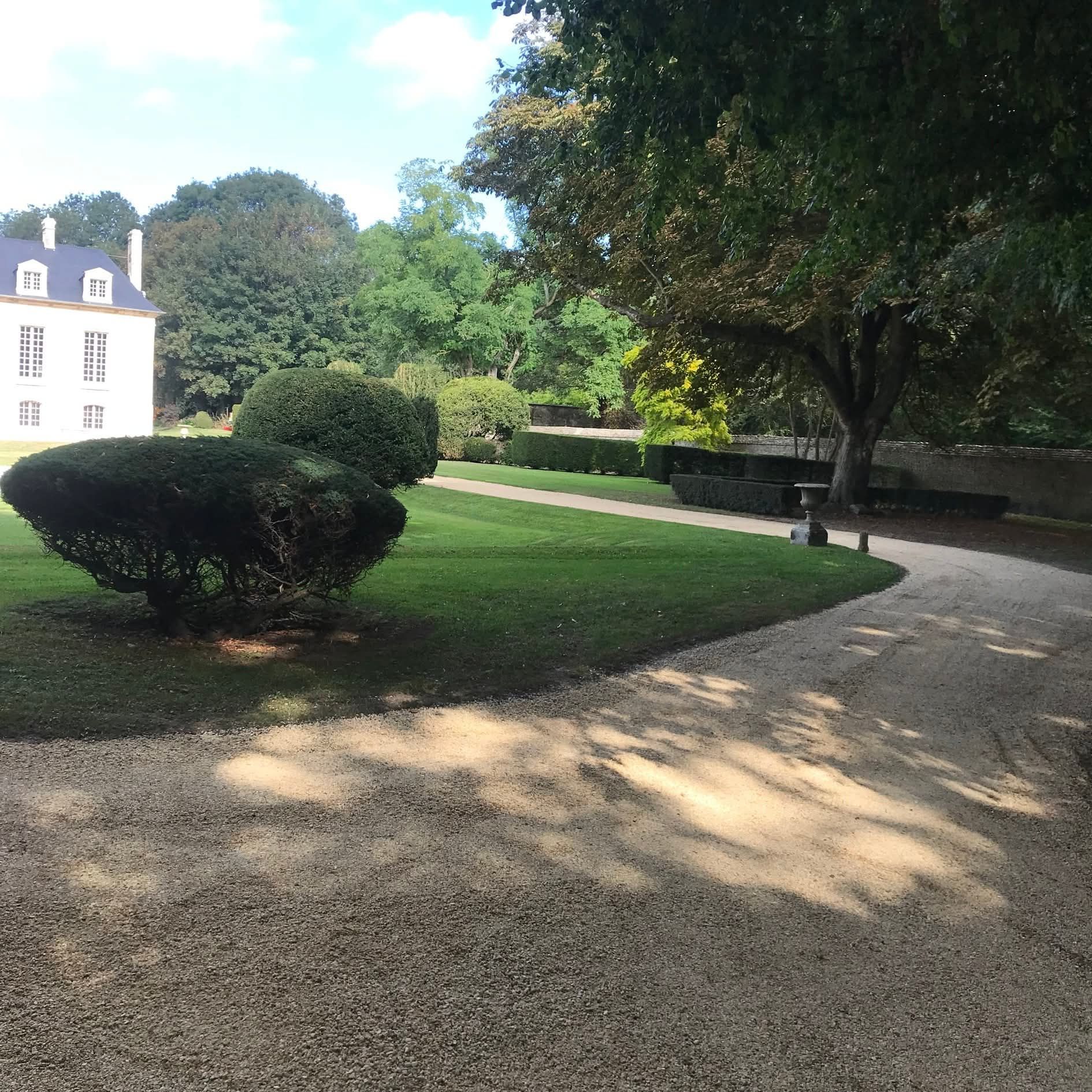 Un chemin de gravier serpente à travers une pelouse bien entretenue en direction d'un bâtiment blanc avec des arbres en arrière-plan.