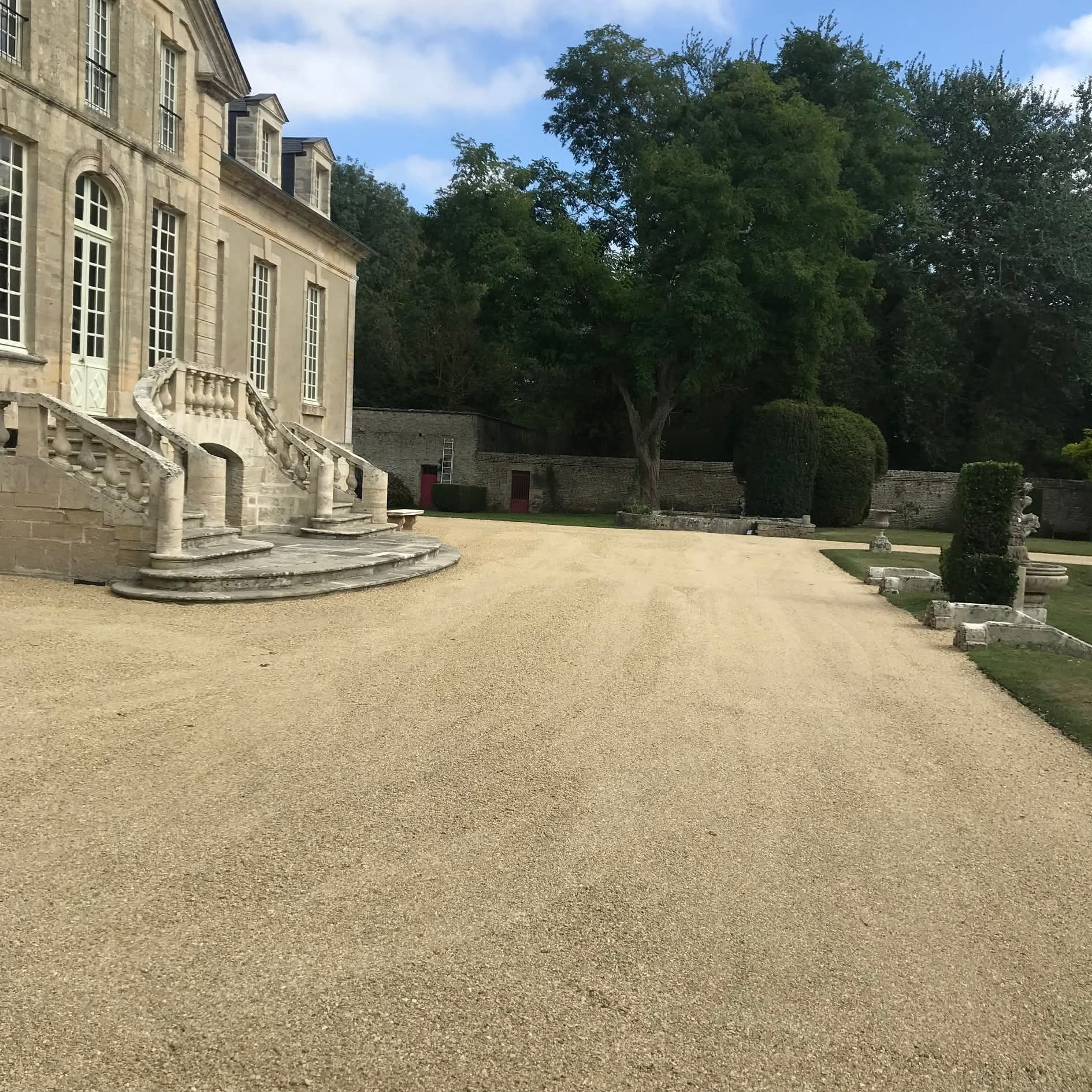 Allée de gravier menant à un château en pierre avec un grand escalier et des haies taillées au cordeau, sous un ciel nuageux.
