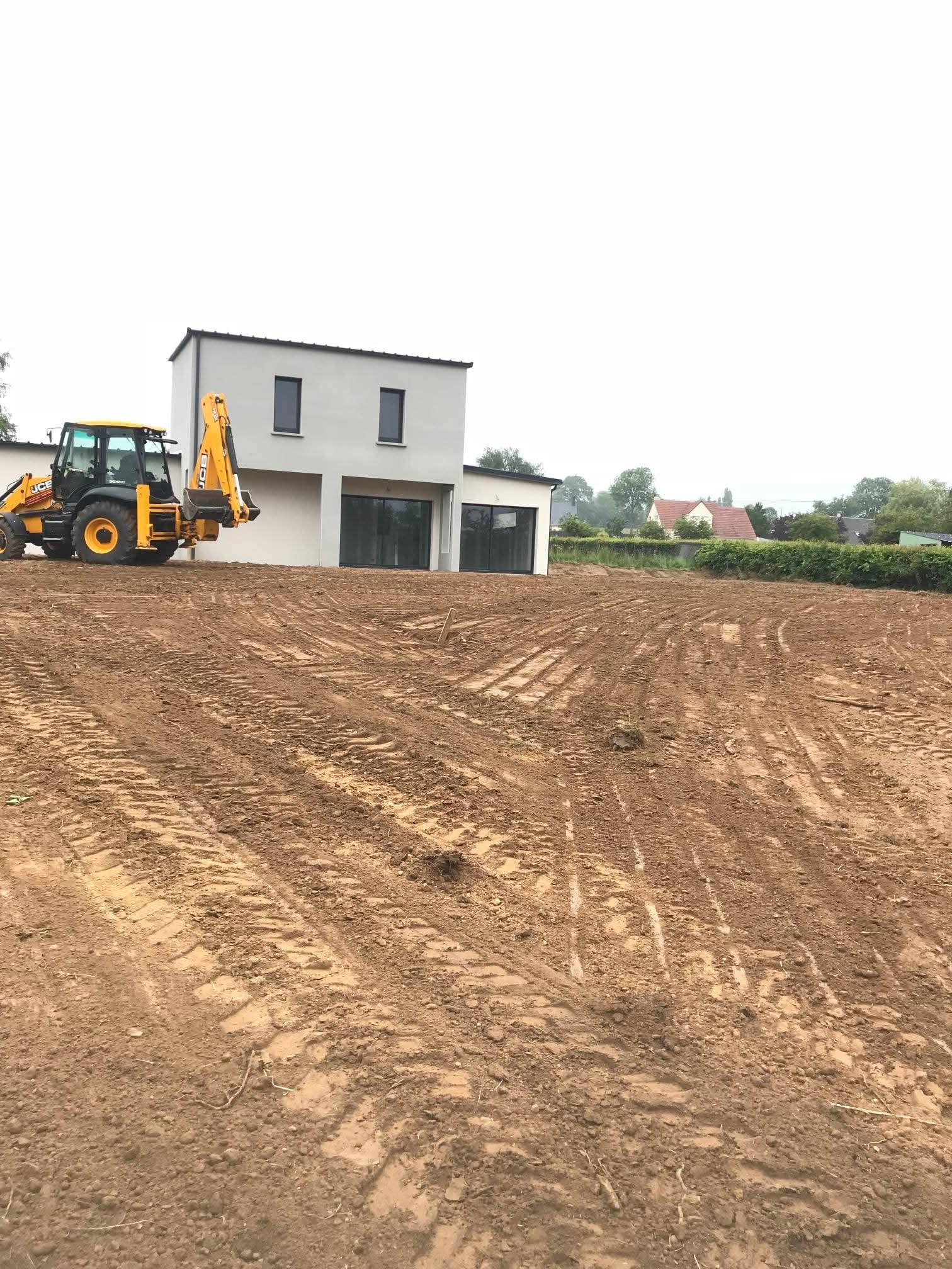 Une pelleteuse sur un terrain vague devant une maison moderne, sous un ciel nuageux.