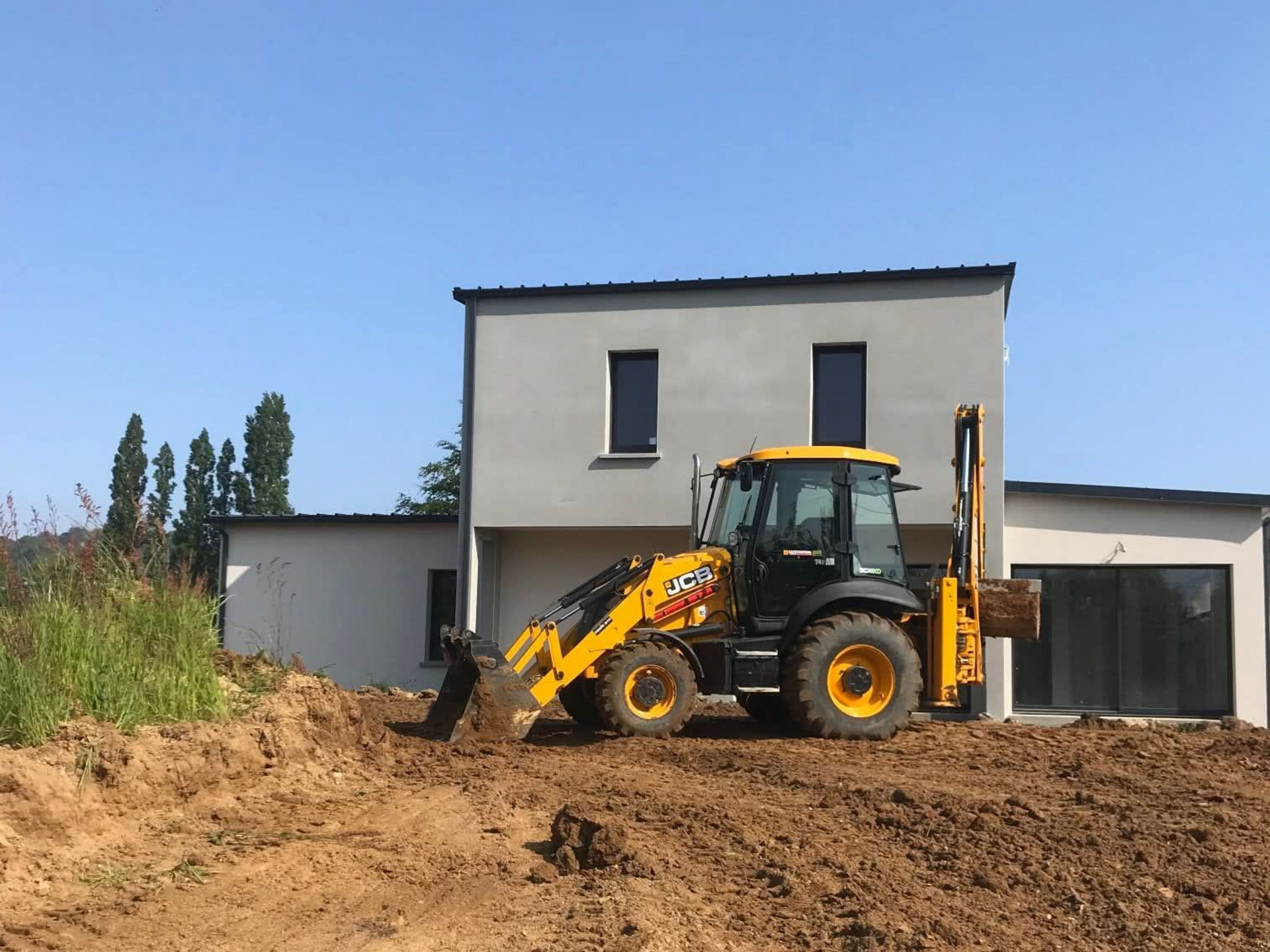 Une pelleteuse creuse la terre près d'une maison moderne et grise, sous un ciel bleu.