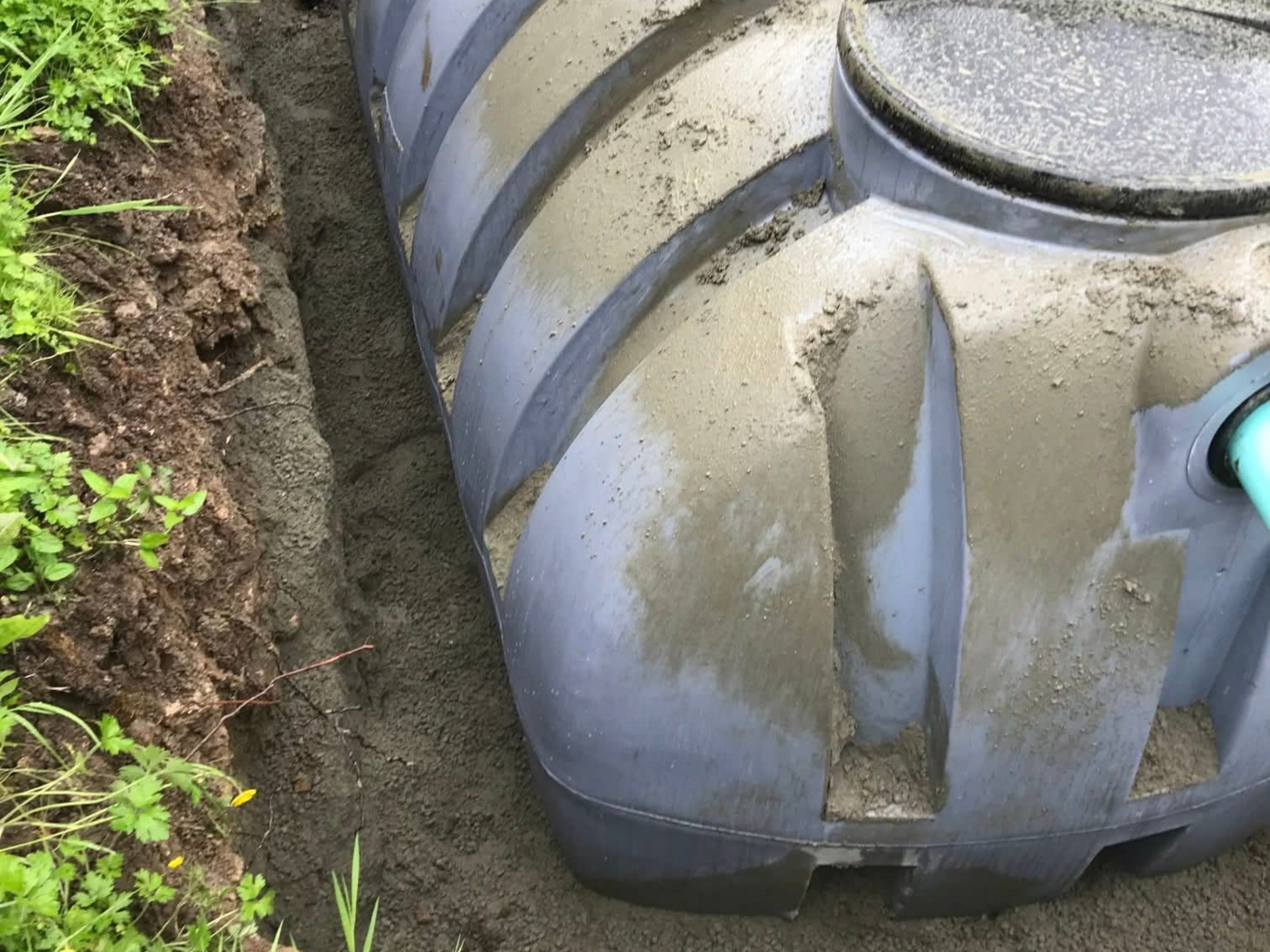 Réservoir en plastique partiellement enterré dans une tranchée, laissant apparaître la terre et la végétation verte.