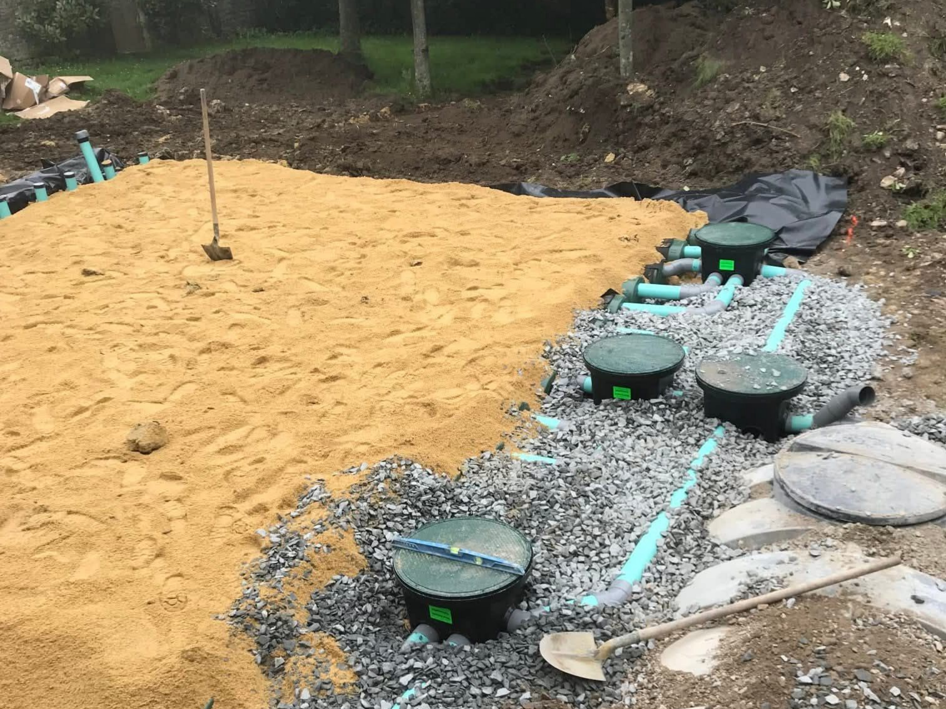 Chantier avec du sable, du gravier et des canalisations. Quatre structures aux toits verts sont visibles, avec une pelle et un paysage en arrière-plan.