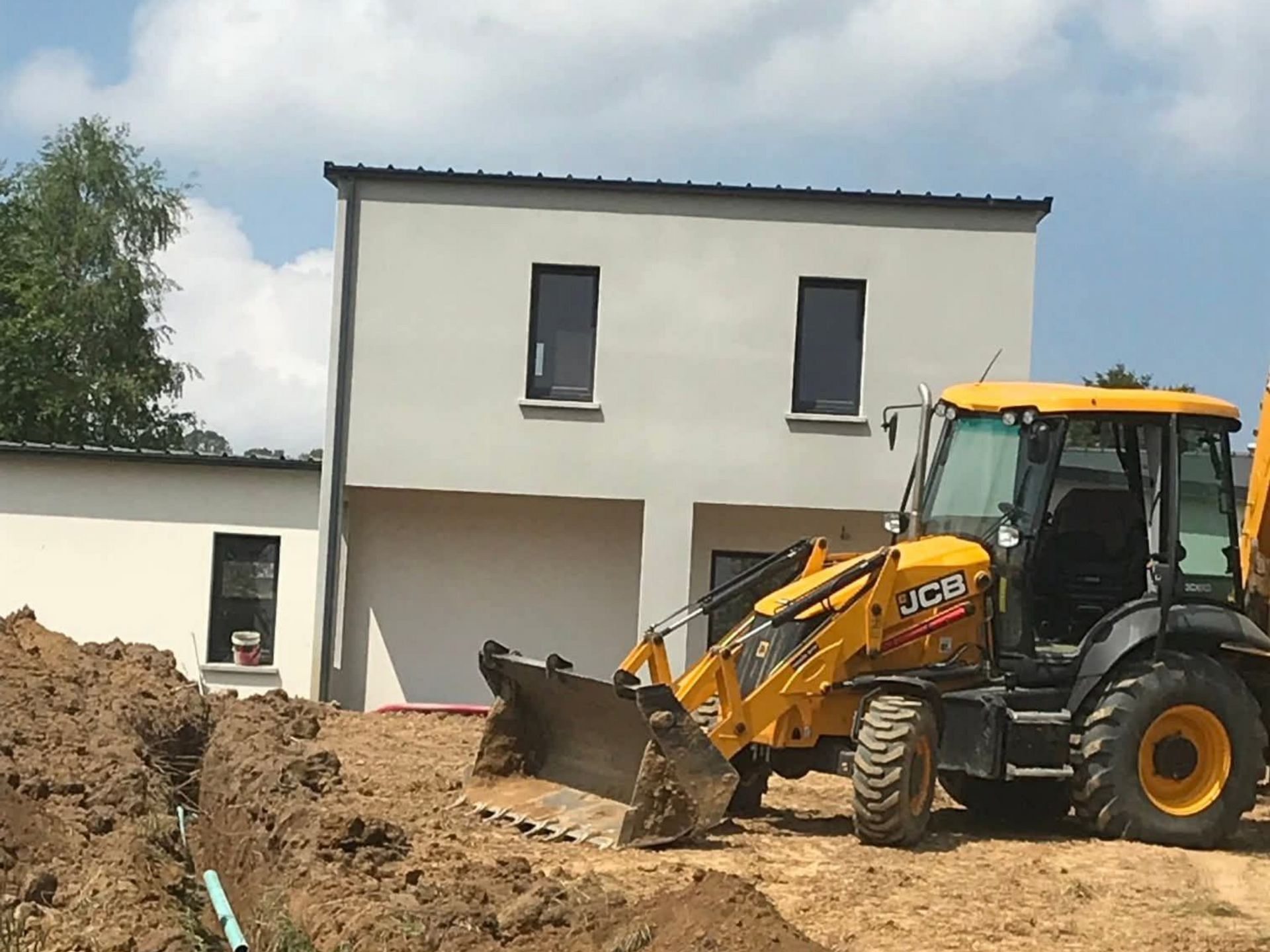 Une pelleteuse jaune creuse près d'un bâtiment gris de deux étages par une journée ensoleillée.