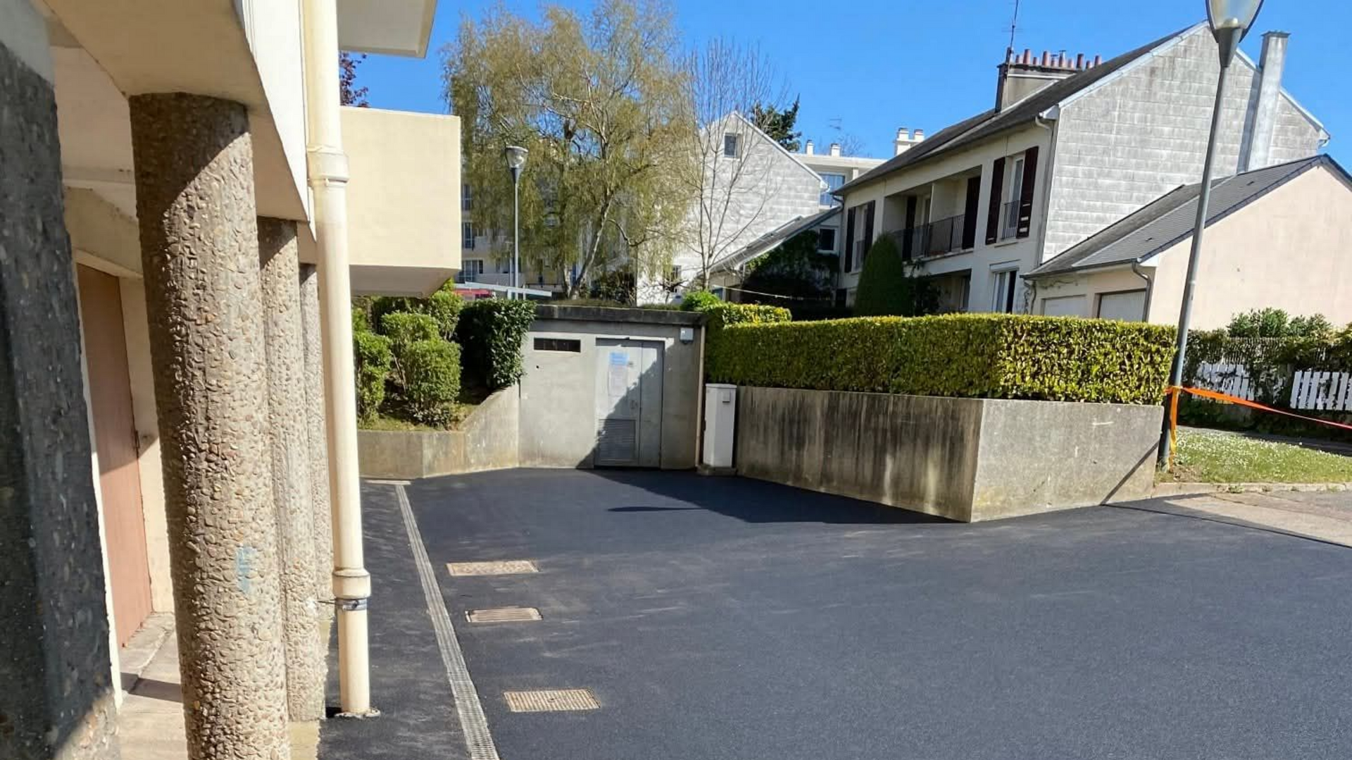 Un parking goudronné devant des bâtiments. Des piliers en pierre à gauche, une haie et un garage à l'arrière-plan.