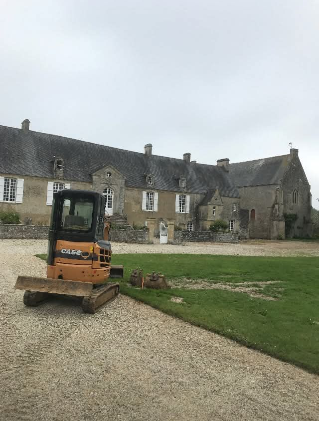 Une pelleteuse sur du gravier devant un grand bâtiment en pierre au toit d'ardoise. Ciel couvert.