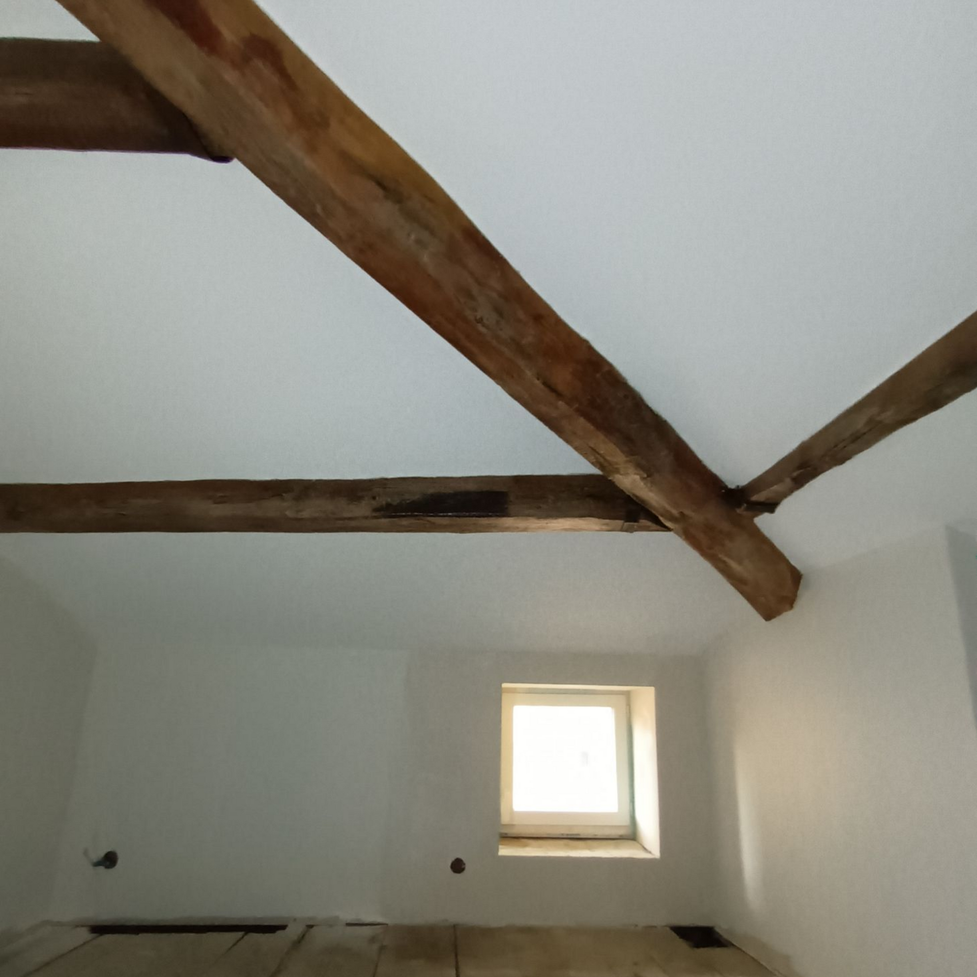 Chambre mansardée avec poutres apparentes et une petite fenêtre carrée sur un mur blanc