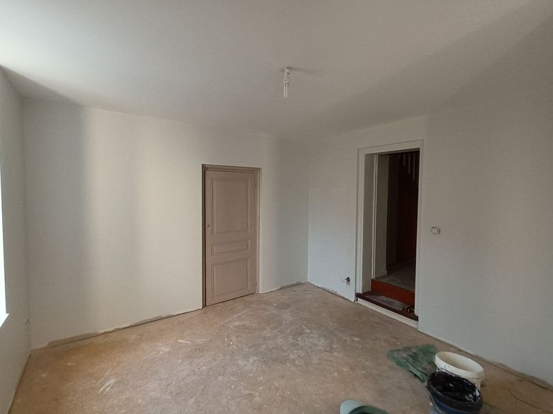 Pièce nue et inachevée, avec des murs blancs, un sol beige et deux portes, dont une donnant sur une zone carrelée de rouge.