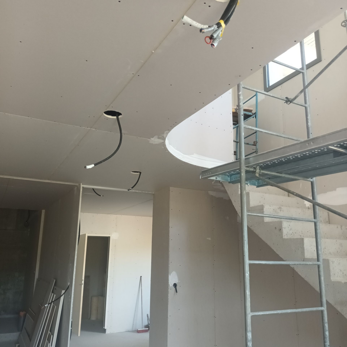 Intérieur en travaux avec cloisons sèches, câblage apparent et échafaudages près d'une cage d'escalier.