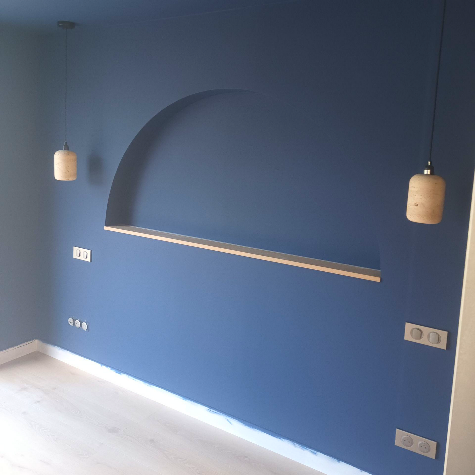 Mur de la pièce peint en bleu avec une niche cintrée en retrait, des suspensions lumineuses et des prises électriques près du sol.