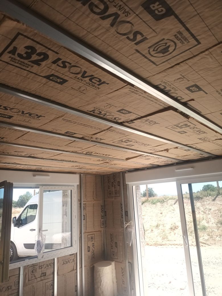 Intérieur d'une pièce inachevée avec des panneaux isolants sur les murs et le plafond, et une fenêtre donnant sur l'extérieur.