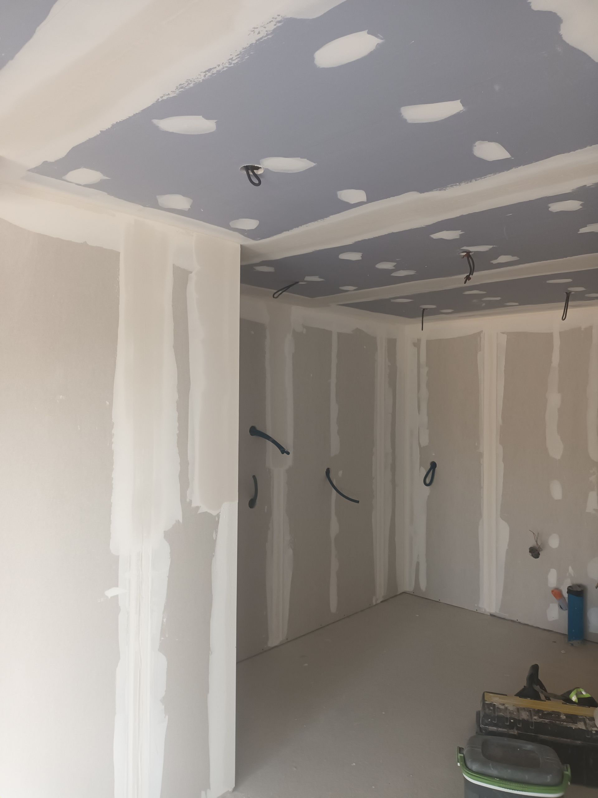 Pièce aux cloisons sèches inachevées, joints collés, plafond gris, fils électriques pendants et outils au sol.