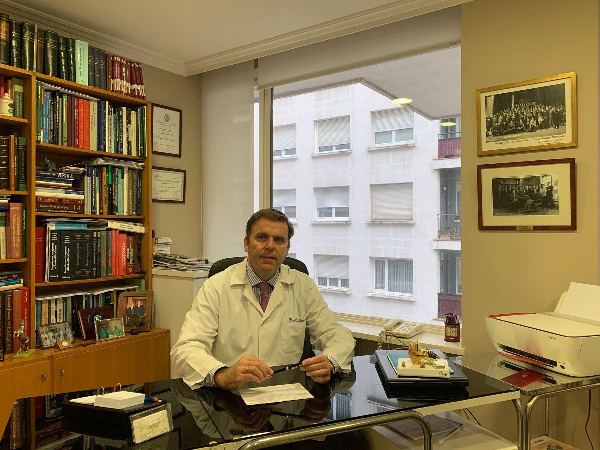 Un hombre con bata de laboratorio se sienta en un escritorio en una oficina, con estanterías y vistas a la ventana del exterior del edificio.