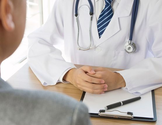 Médico con bata blanca y estetoscopio, consultando con un paciente en un escritorio, con bloc de notas y bolígrafo presentes.