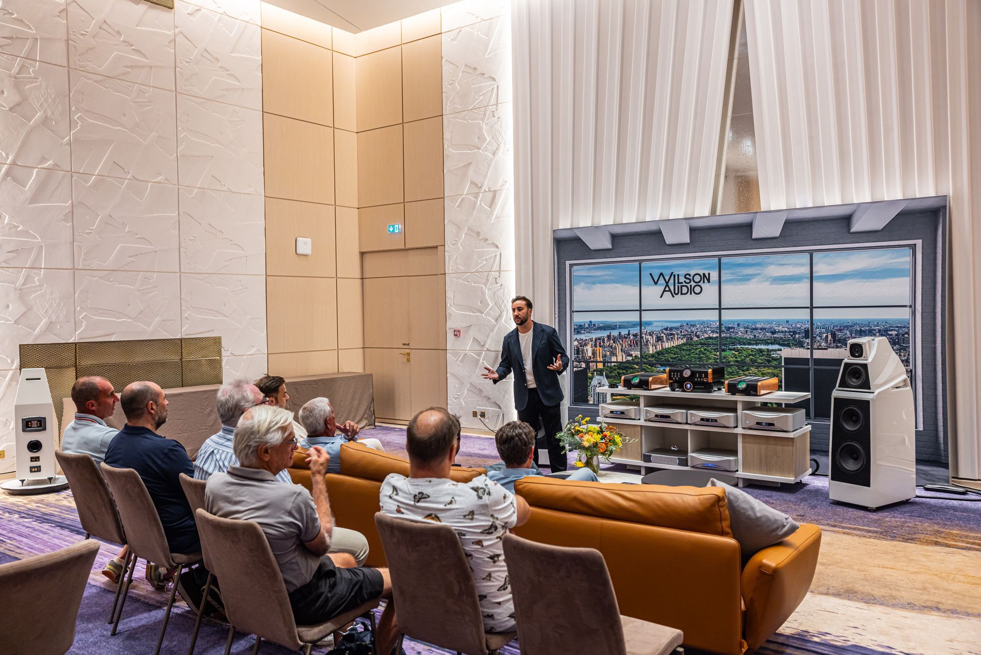 Un homme présente du matériel audio à un public assis dans une pièce lumineuse.