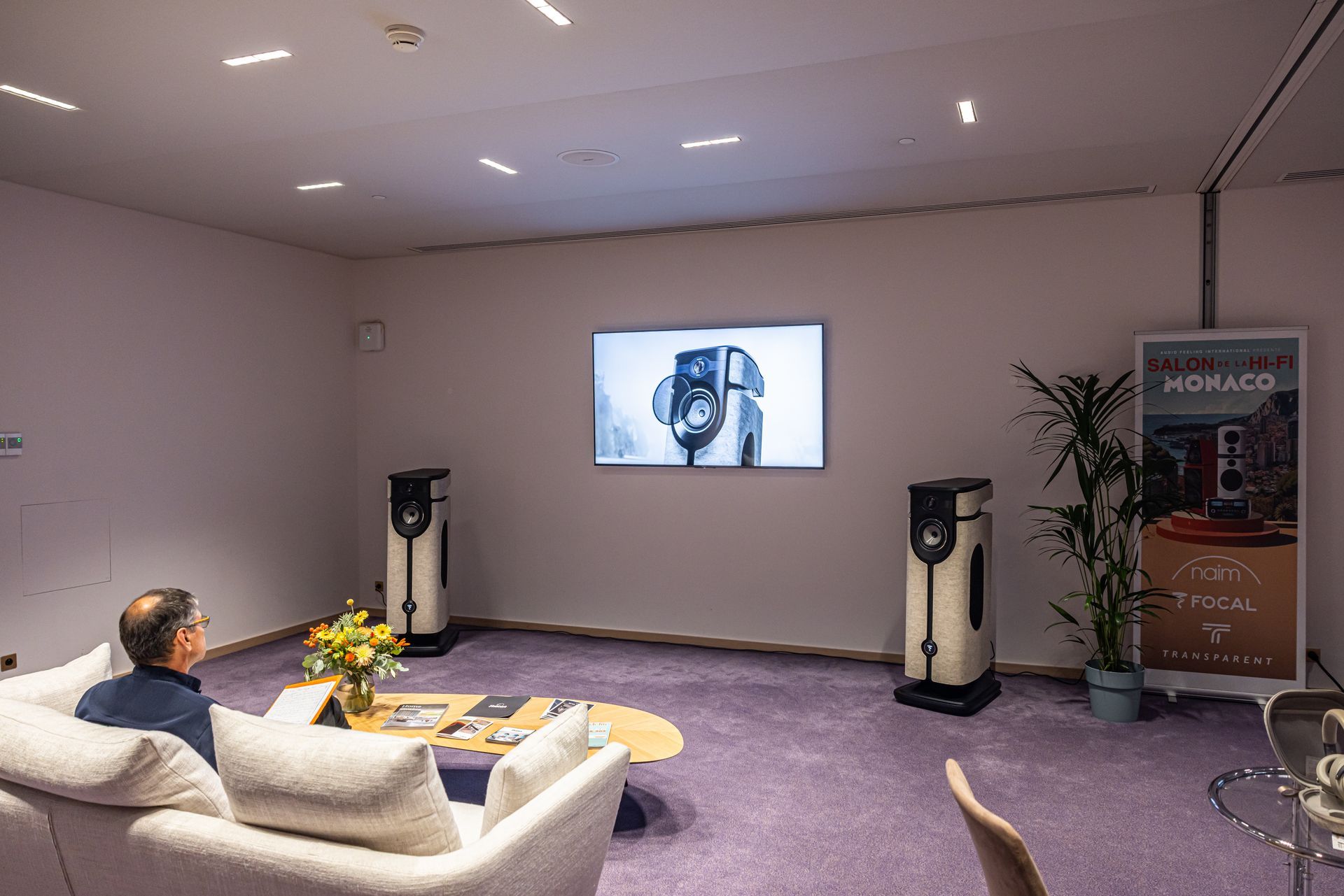 Un homme est assis sur un canapé, regardant un écran dans une pièce moderne équipée de haut-parleurs et de supports promotionnels.