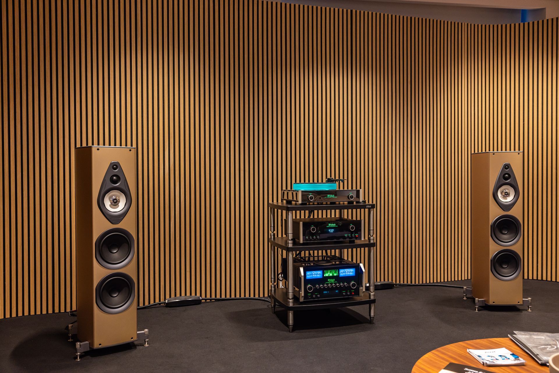 Deux enceintes beiges encadrent un équipement audio posé sur un support, contre un mur à lattes de bois.