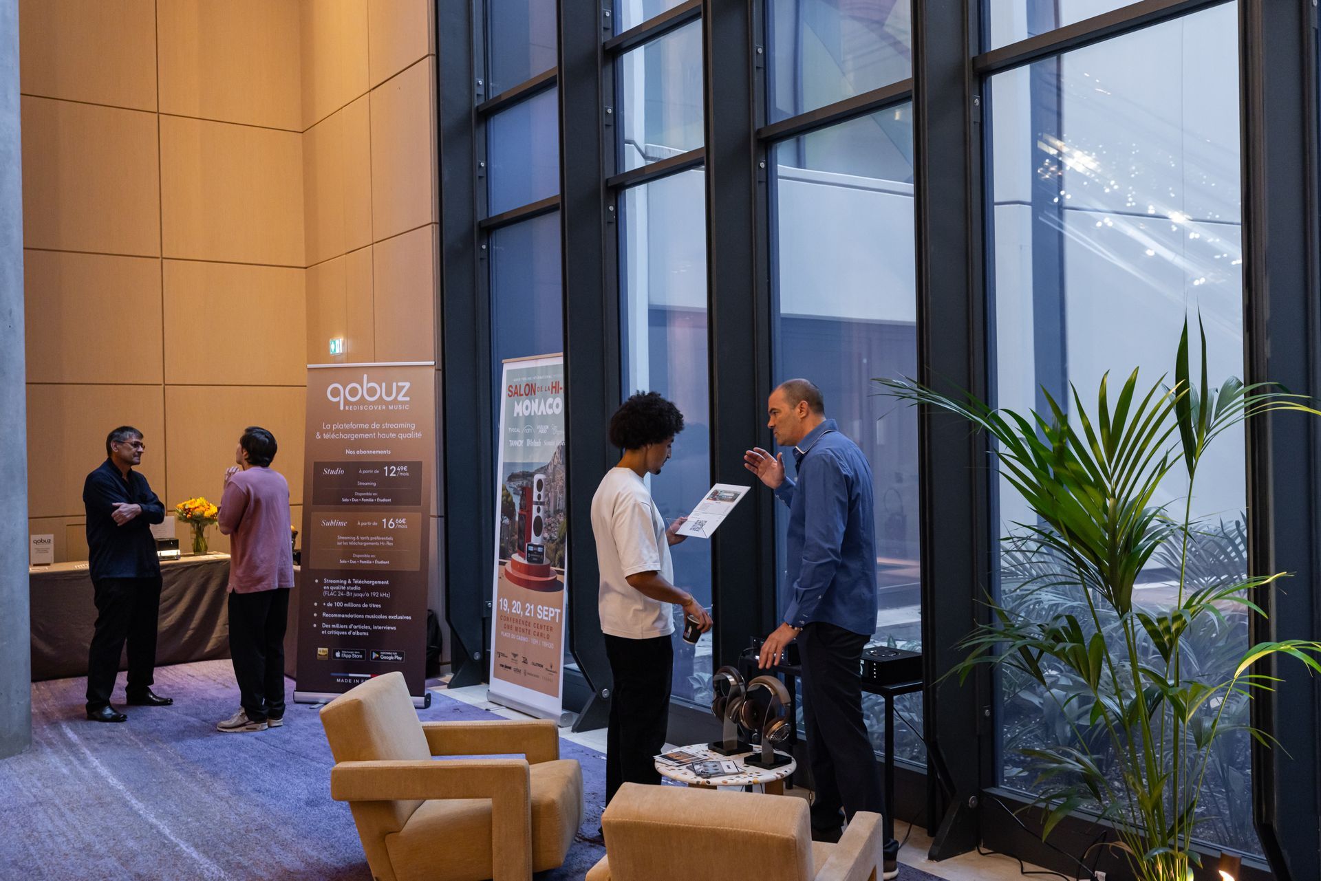 Des personnes discutent près de stands ornés de bannières publicitaires lors d'un événement. De hautes fenêtres et des plantes se dessinent en arrière-plan.