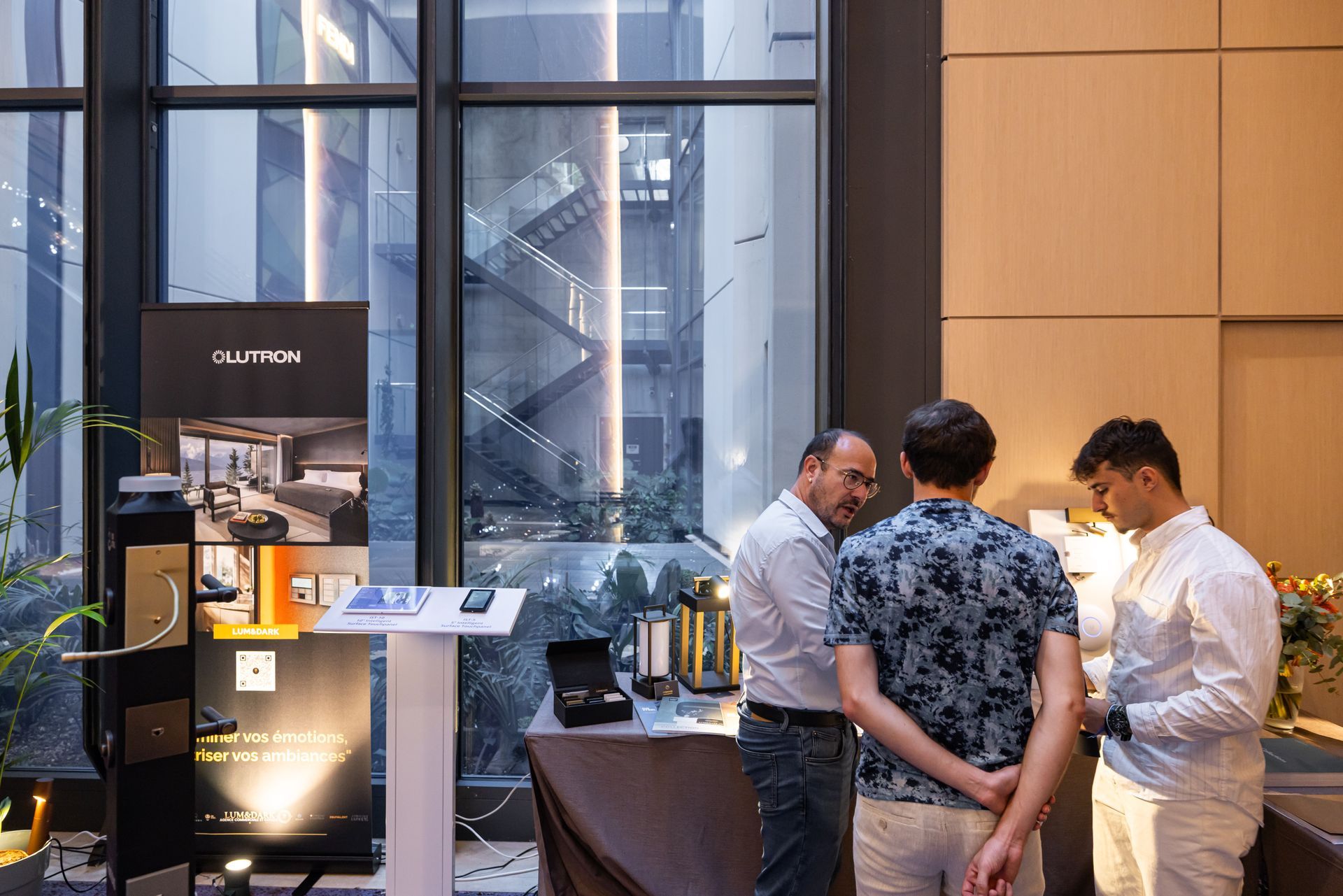 Des personnes examinent des produits exposés sur une table lors d'un événement, à l'intérieur d'un bâtiment doté de grandes fenêtres.