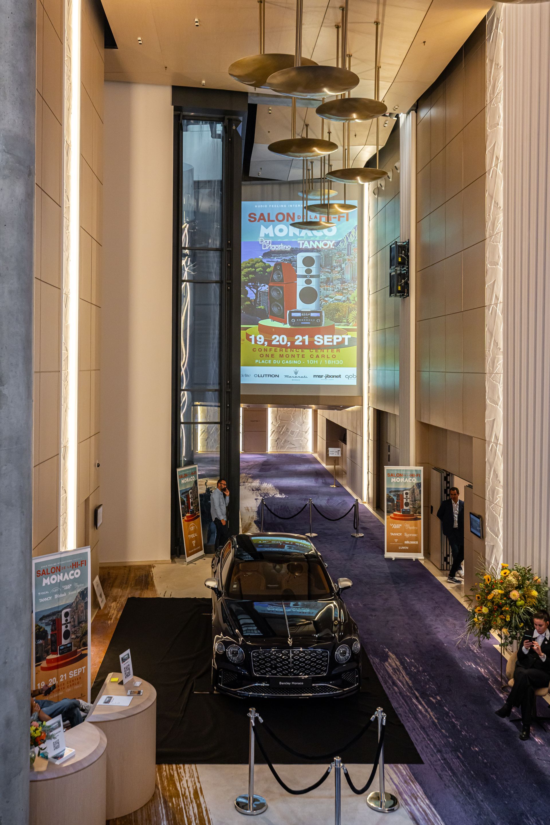 Hall d'entrée avec voiture noire exposée, affiche de l'événement, moquette et éclairage décoratif.